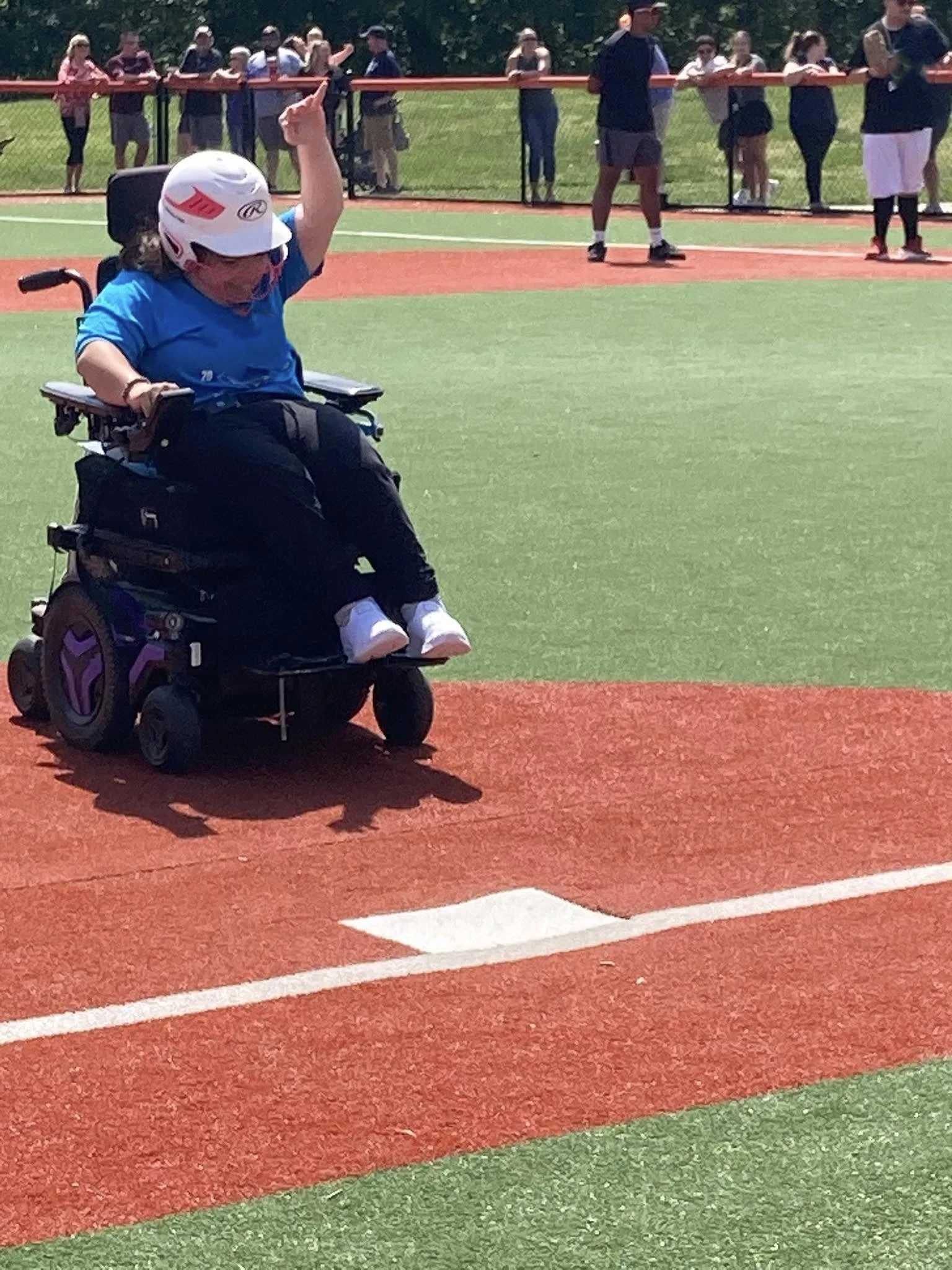 A person in a wheelchair wearing a helmet and blue shirt, celebrating or cheering at a sports event on a field with a group of people in the background.