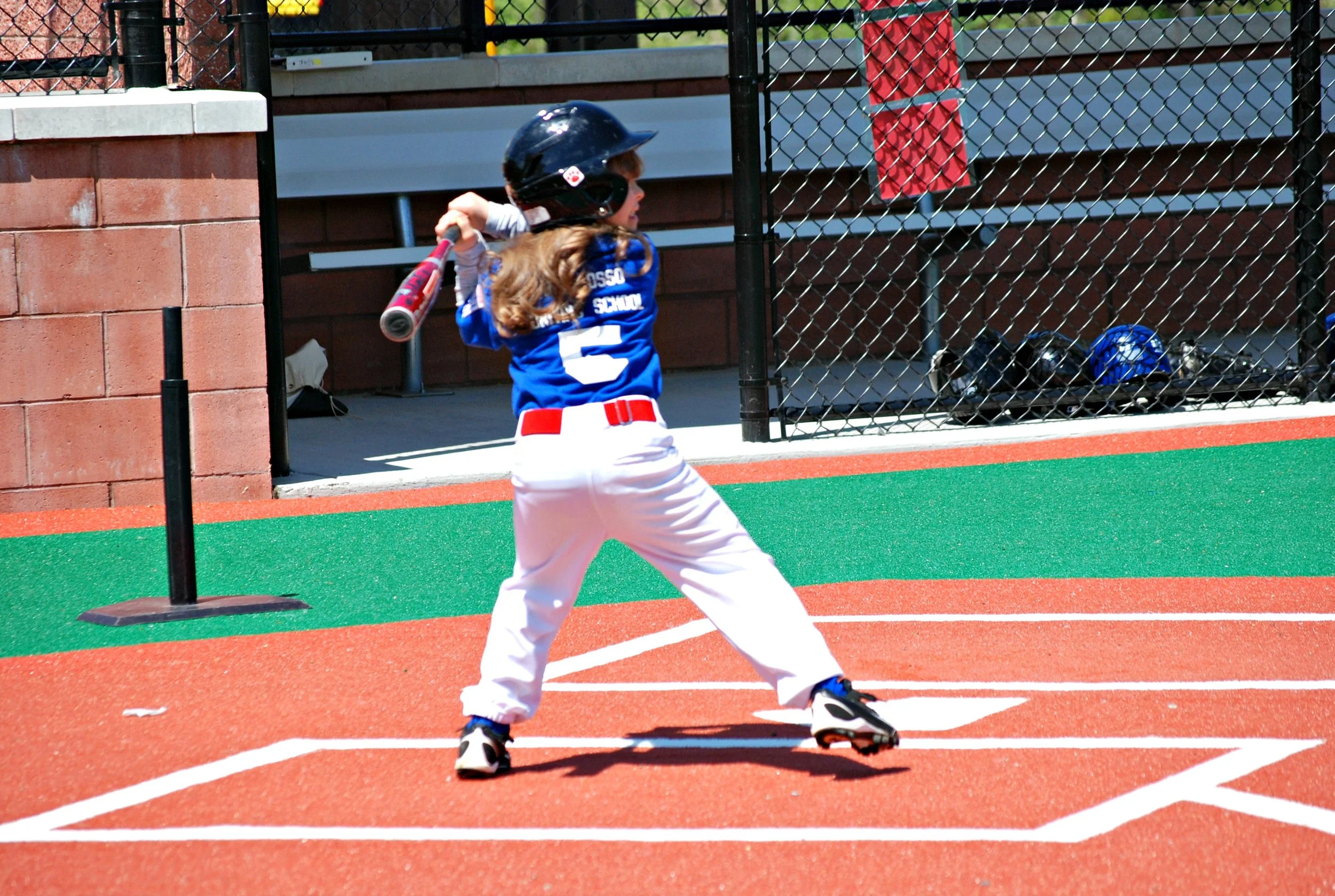 A young girl in a baseball uniform and helmet swinging a bat at the baseball field.