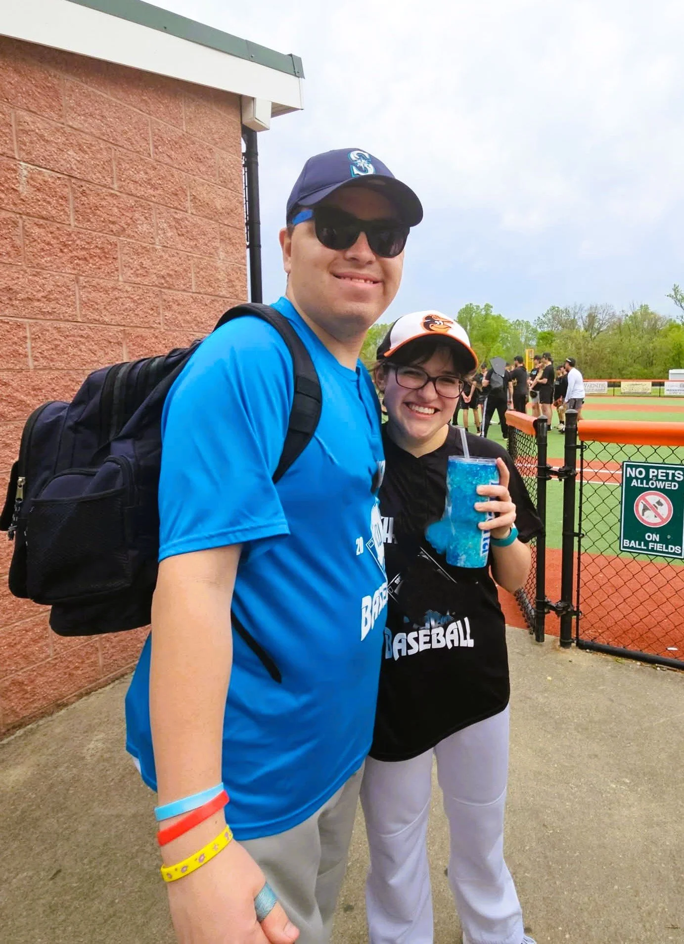 A man and a young girl at a baseball game. The man is wearing sunglasses, a blue shirt, and a cap, carrying a black backpack. The girl is holding a blue drink and wearing glasses, a black jersey, and a baseball cap. They are standing near a fenced ba
