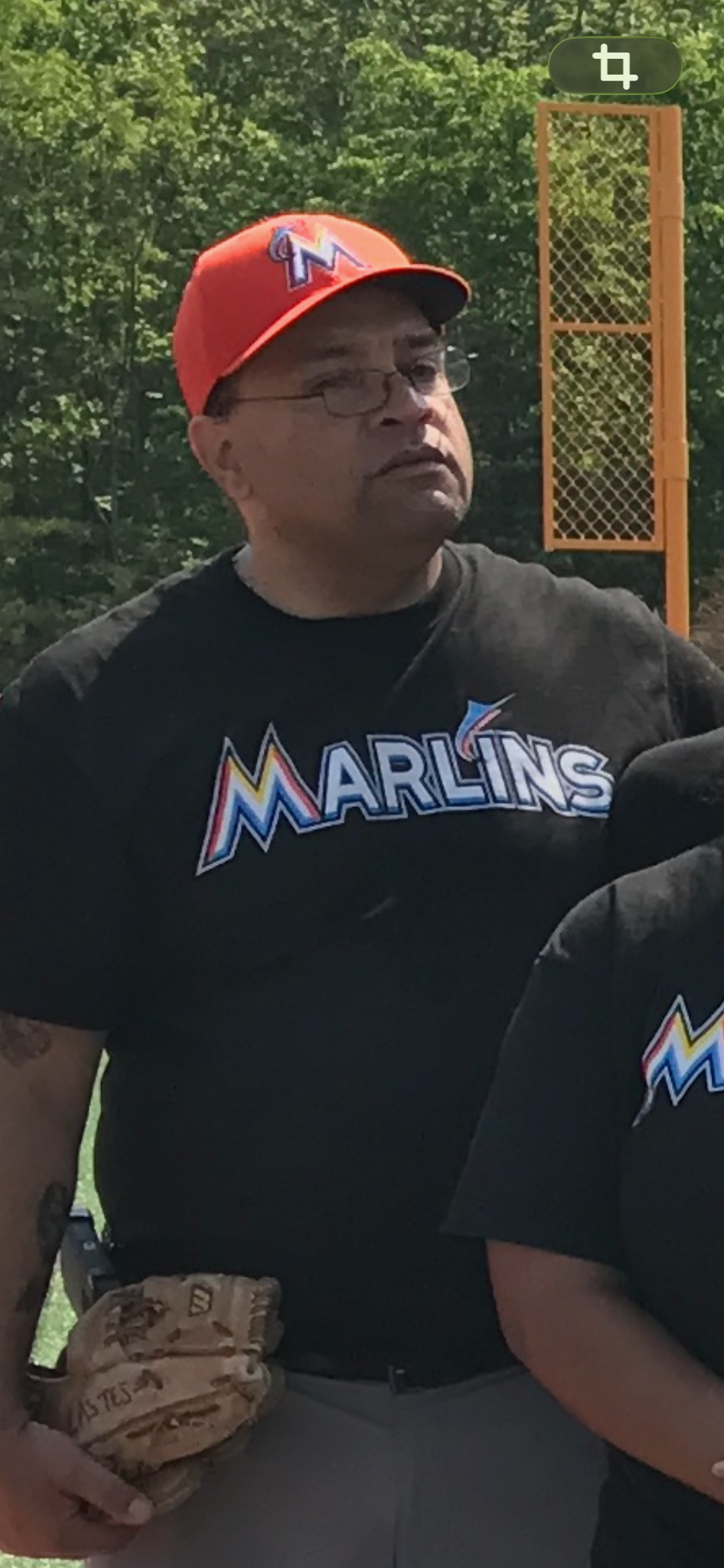 A man wearing glasses, a red Miami Marlins baseball cap, and a black Marlins t-shirt stands outdoors with greenery in the background, near a chain-link backstop.