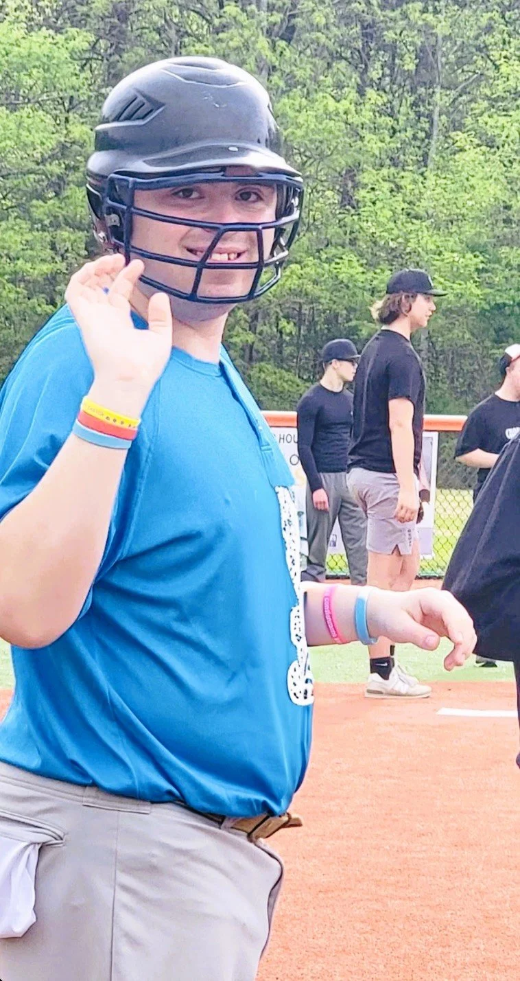 A young person wearing a baseball helmet and blue t-shirt, smiling and waving, at a baseball field, with others in the background.