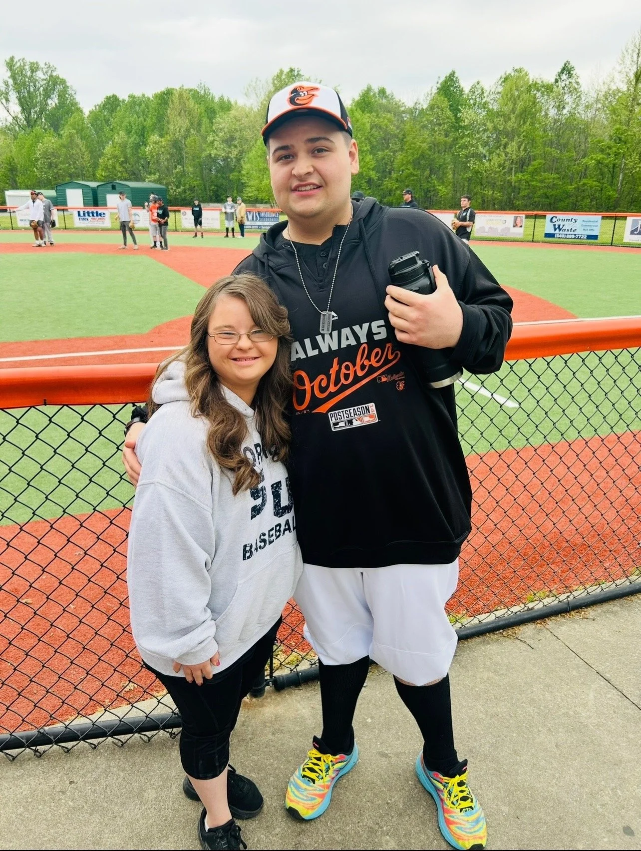 A young girl and a baseball player smiling and posing together at a baseball field. The girl is wearing glasses, a gray hoodie, and black pants. The baseball player is wearing a Baltimore Orioles hoodie, white baseball pants, colorful sneakers, and a
