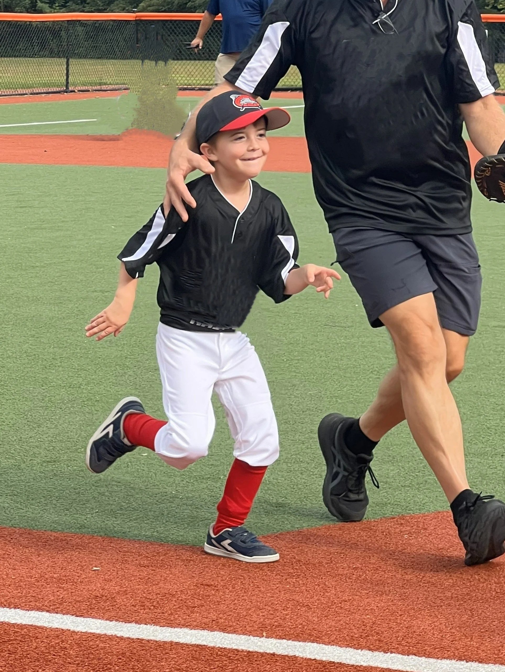 A young boy in a baseball uniform running on a baseball field near an adult coach, who is wearing a black shirt and shorts, during a game or practice.
