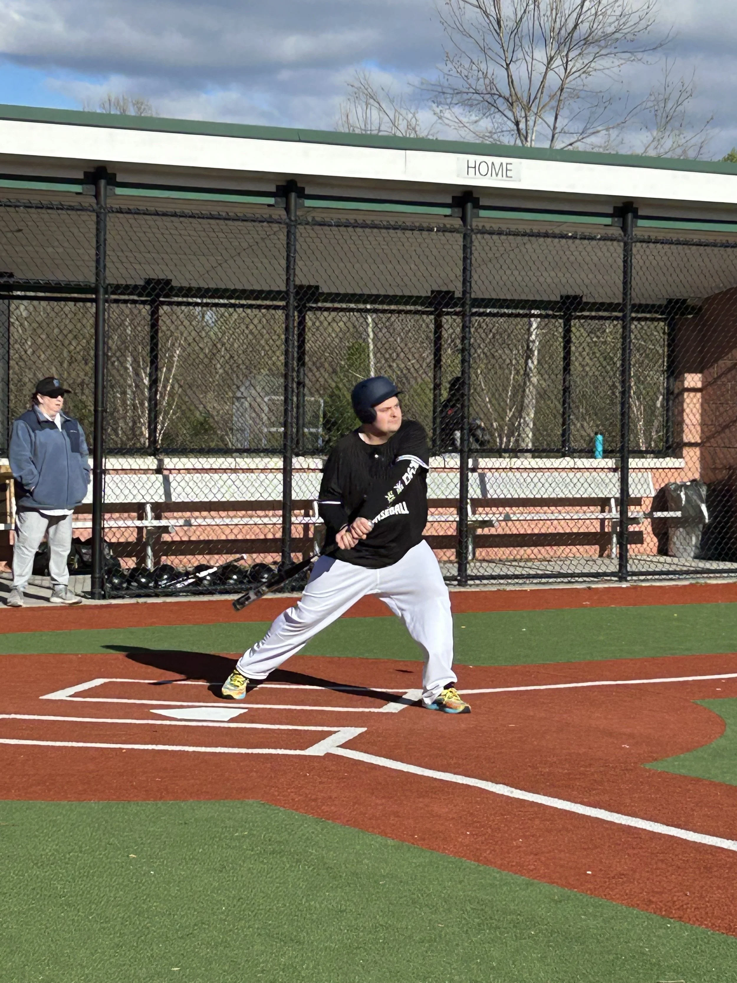 A baseball player in a batting stance at home plate during a game, with a coach and others watching behind a fence.