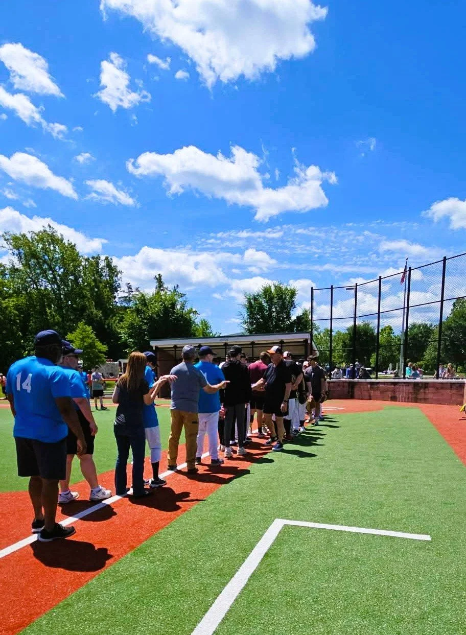 People standing in line on a baseball field under a partly cloudy sky, with trees in the background.
