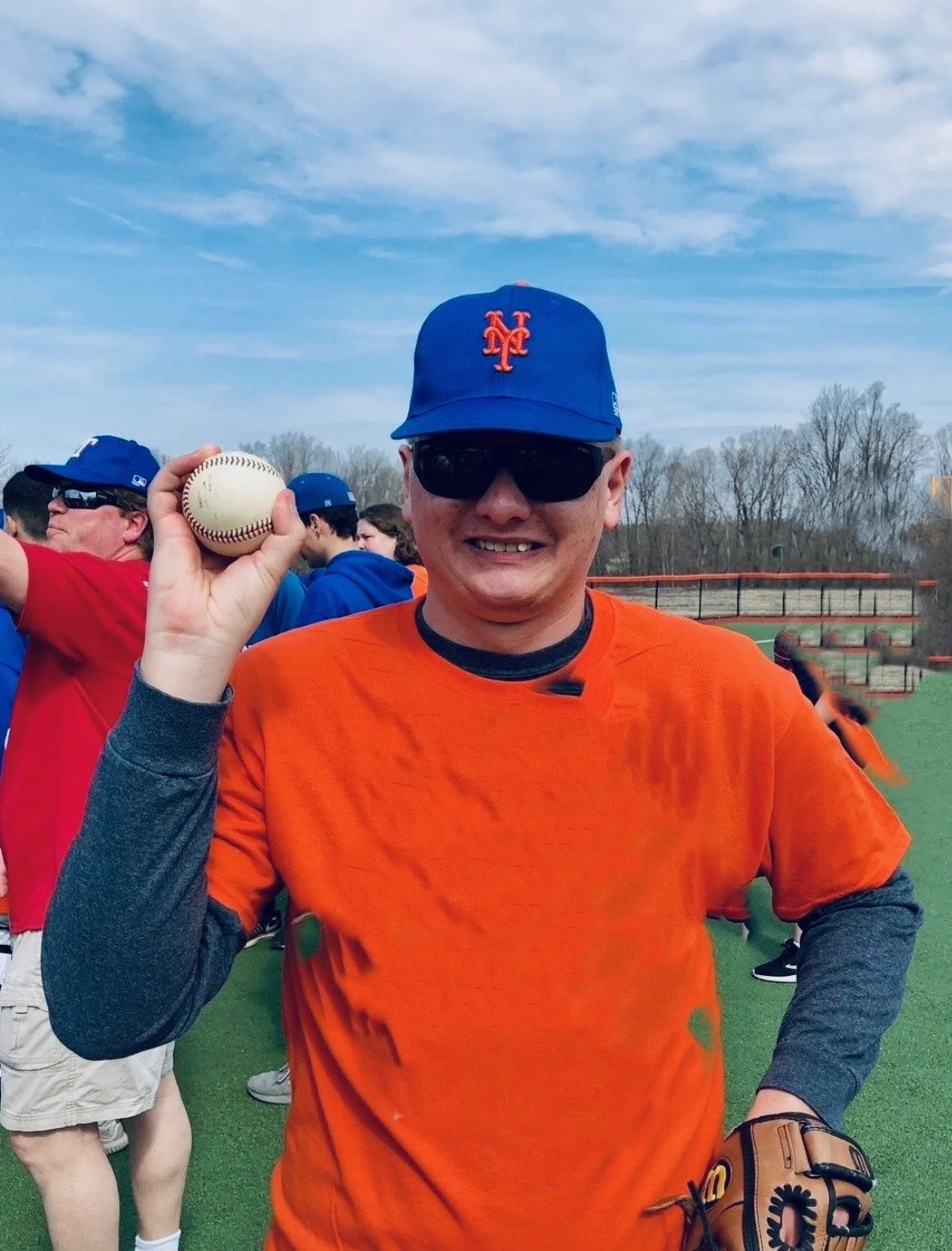 A man in sunglasses, a blue baseball cap with an orange logo, and an orange shirt is holding a baseball. He is smiling on a baseball field with others in the background.