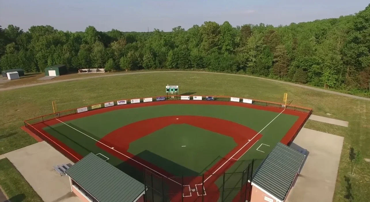 An aerial view of a small professional baseball or softball field with green grass in the outfield and a red infield, surrounded by trees and a grassy area. The field has a seating area with a green roof on the right side and a small scoreboard in center field.