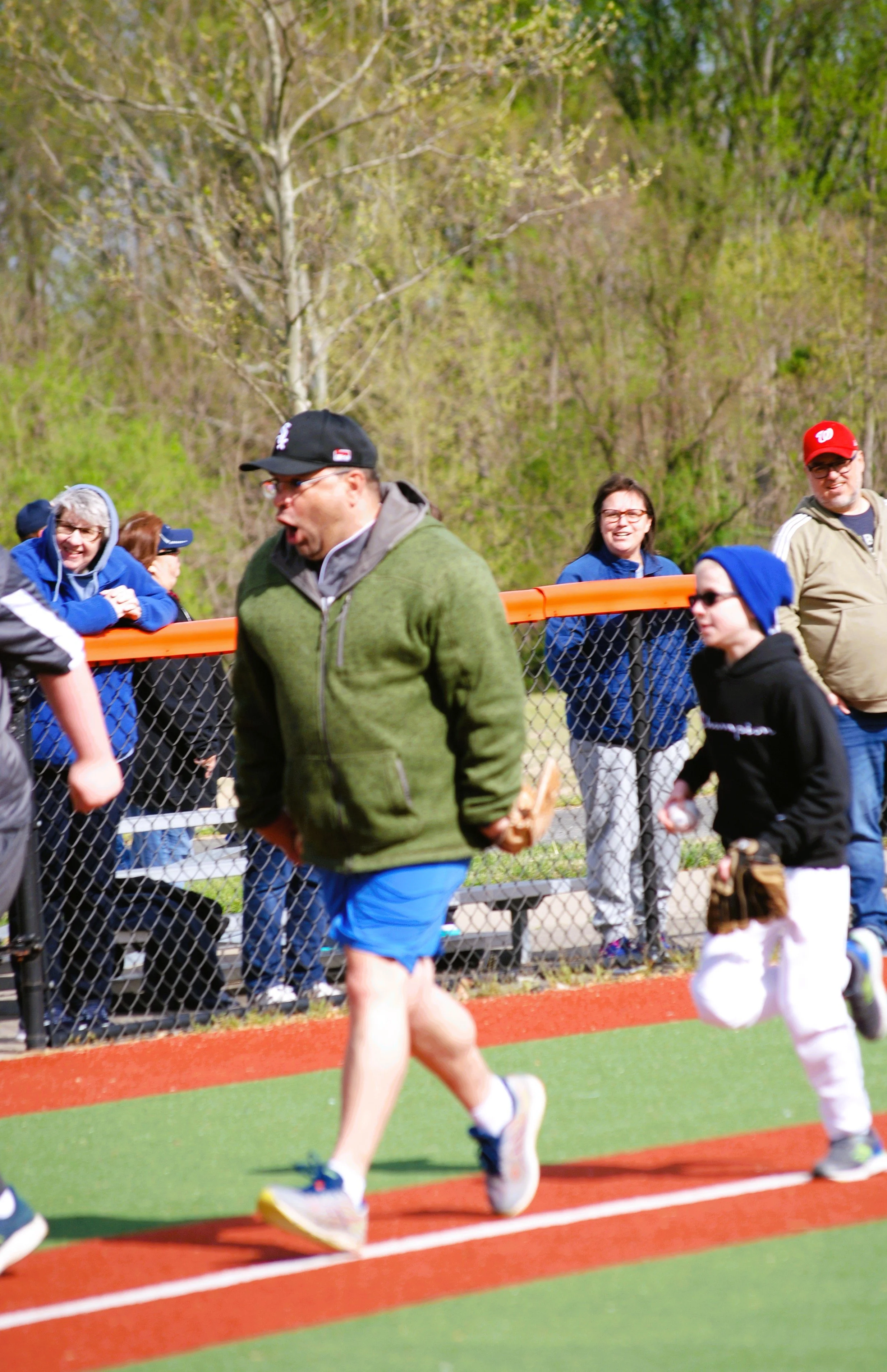 A group of people at a baseball or softball game, with a man in a green jacket and blue shorts, and a boy in a baseball uniform running on the field. Spectators are standing behind a chain-link fence, watching and smiling.