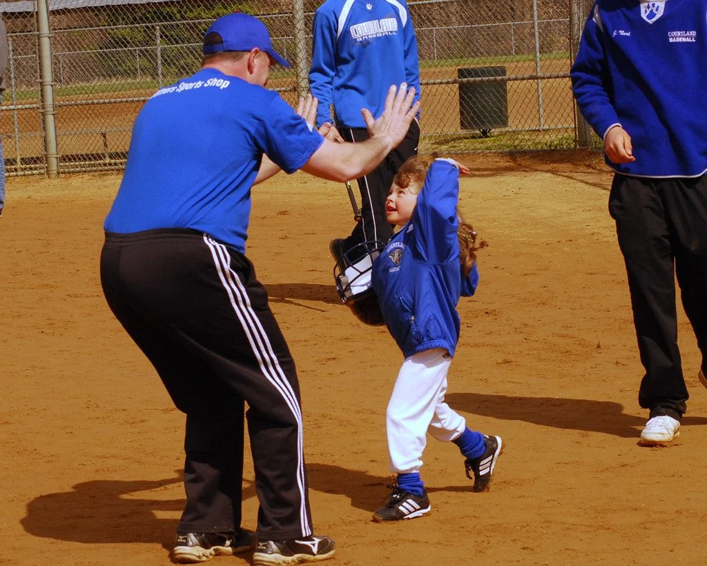 A young girl in a blue jacket and white pants on a baseball field, giving a high five to an adult coach wearing a blue shirt and black pants, with two other coaches nearby.