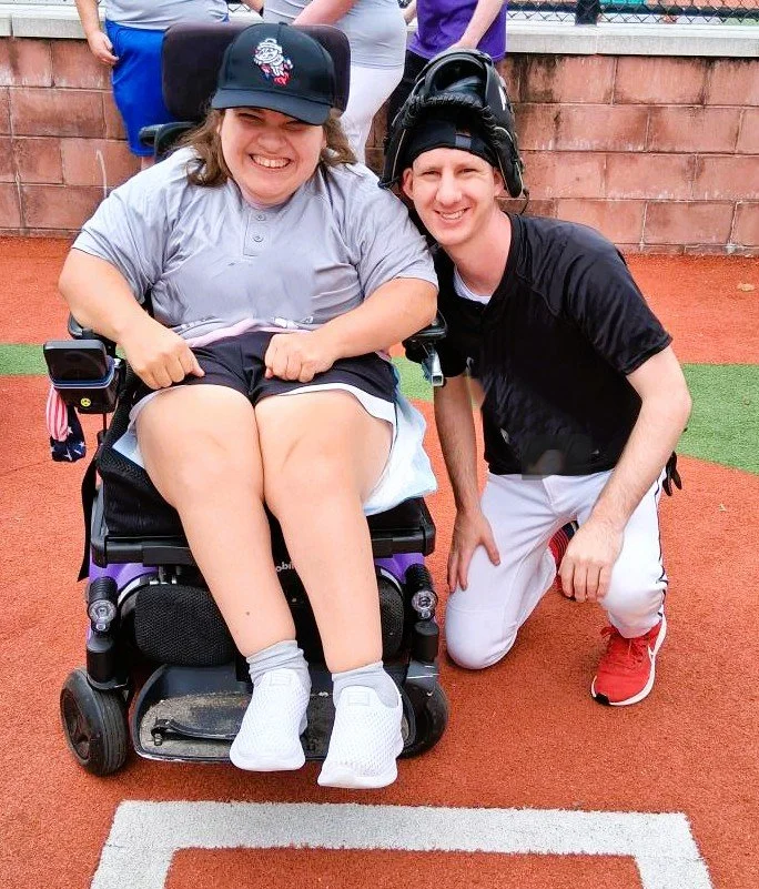 A person in a wheelchair with a baseball cap and shirt, smiling, next to a man in a baseball uniform, kneeling on one knee, both smiling, on a baseball field.