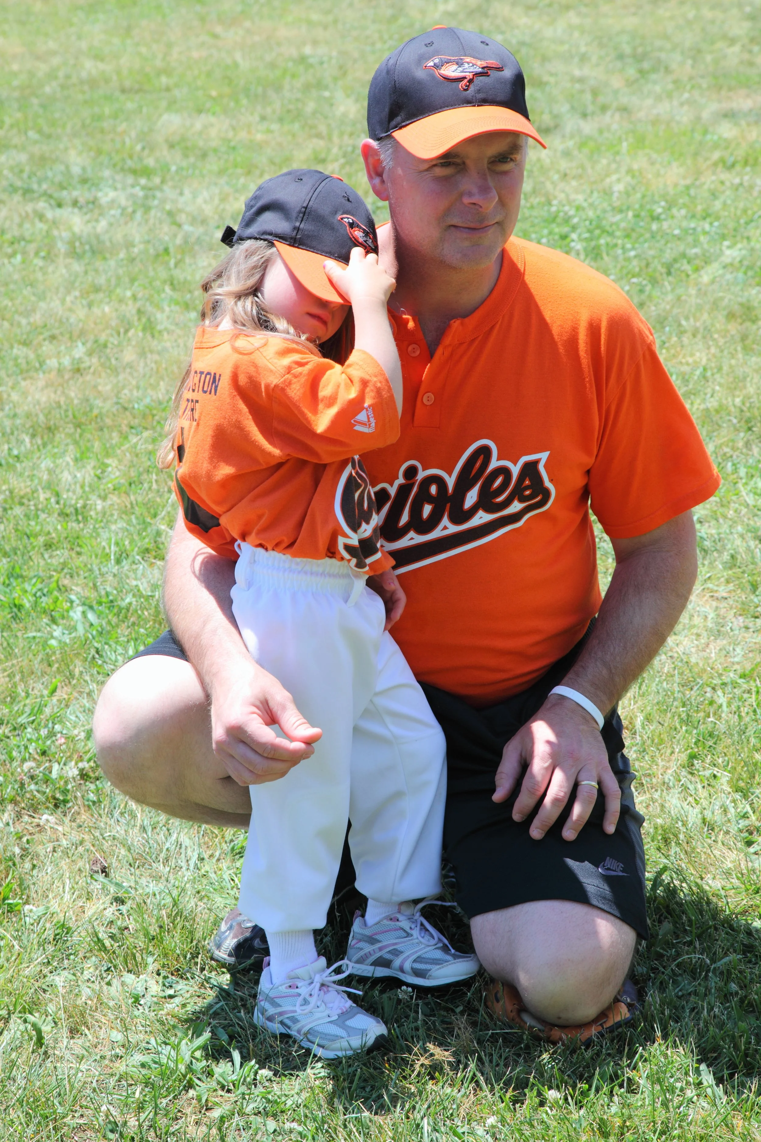 A man kneeling on one knee on the grass with a young girl leaning on his shoulder, both wearing Baltimore Orioles baseball team apparel and hats. The girl appears tired or resting, and both are outdoors on a sunny day.