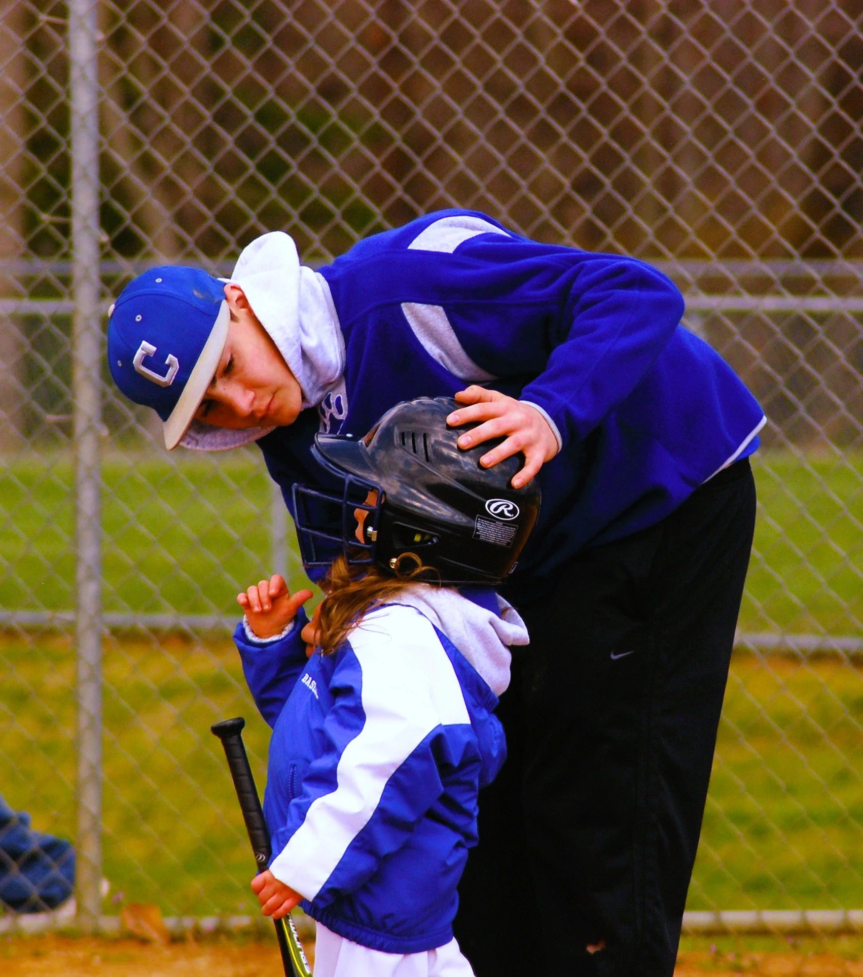 A young boy in a blue baseball uniform and cap leaning down to listen to a coach or older player who is also in a blue uniform, while holding a baseball bat. They appear to be on a baseball field near a chain-link fence.