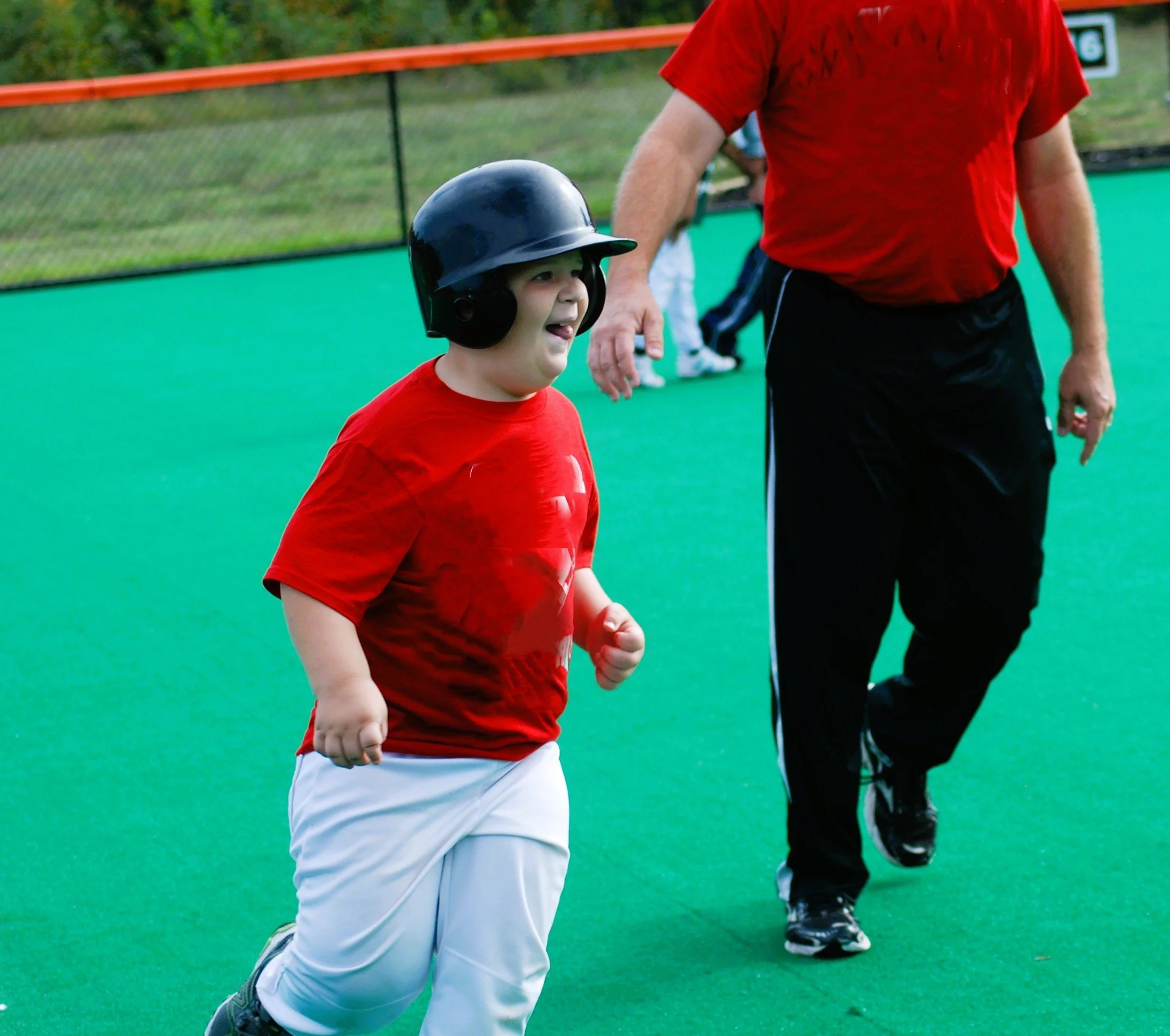 A young boy in a red shirt and white pants wearing a black batting helmet, running on a green softball field with a coach or adult nearby.