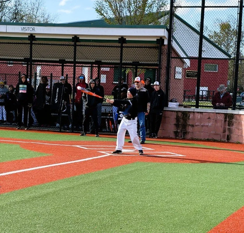 A young baseball player preparing to bat at a baseball field with spectators and team members watching from behind a chain-link fence.