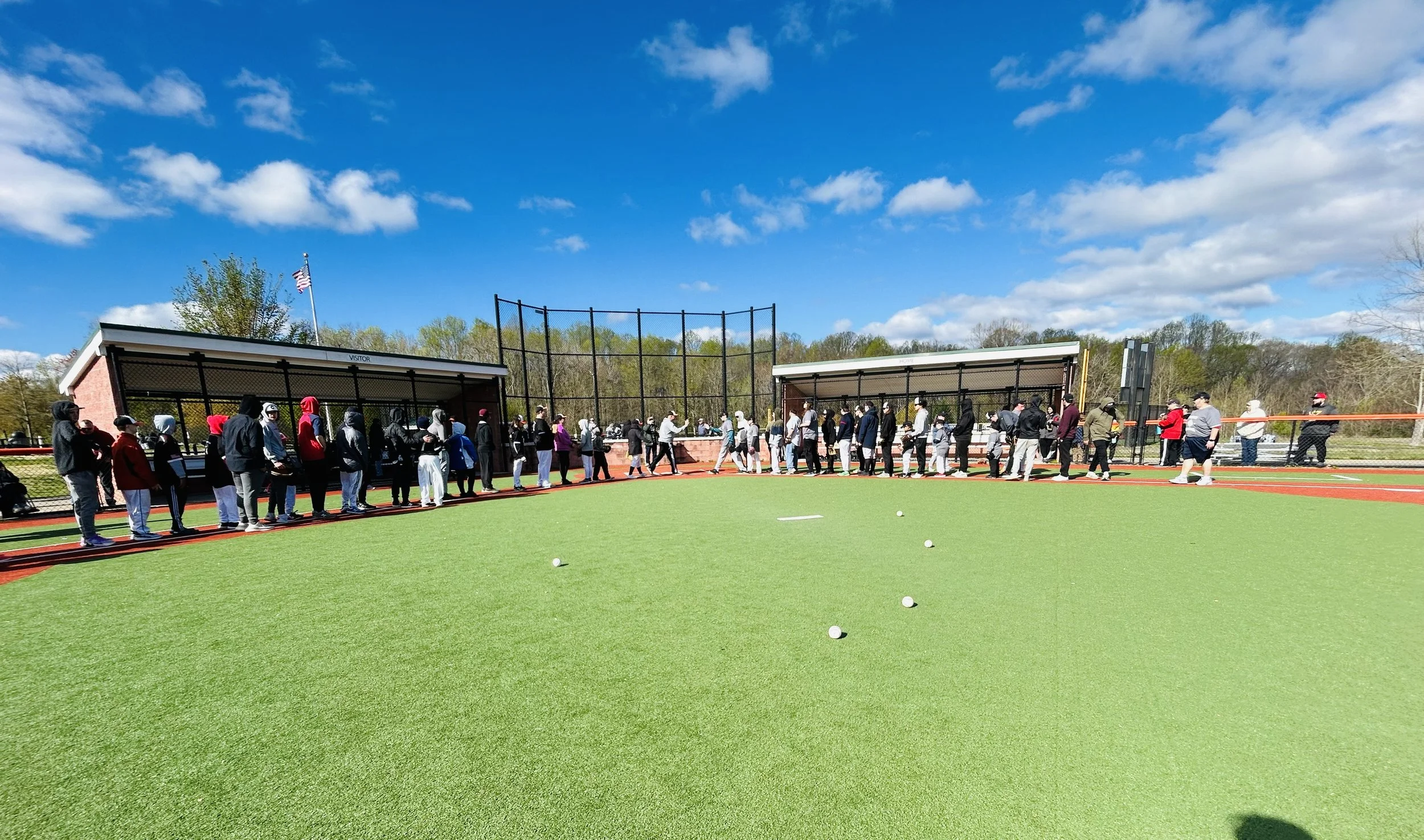 Line of people waiting near a baseball dugout at a sports field on a sunny day.