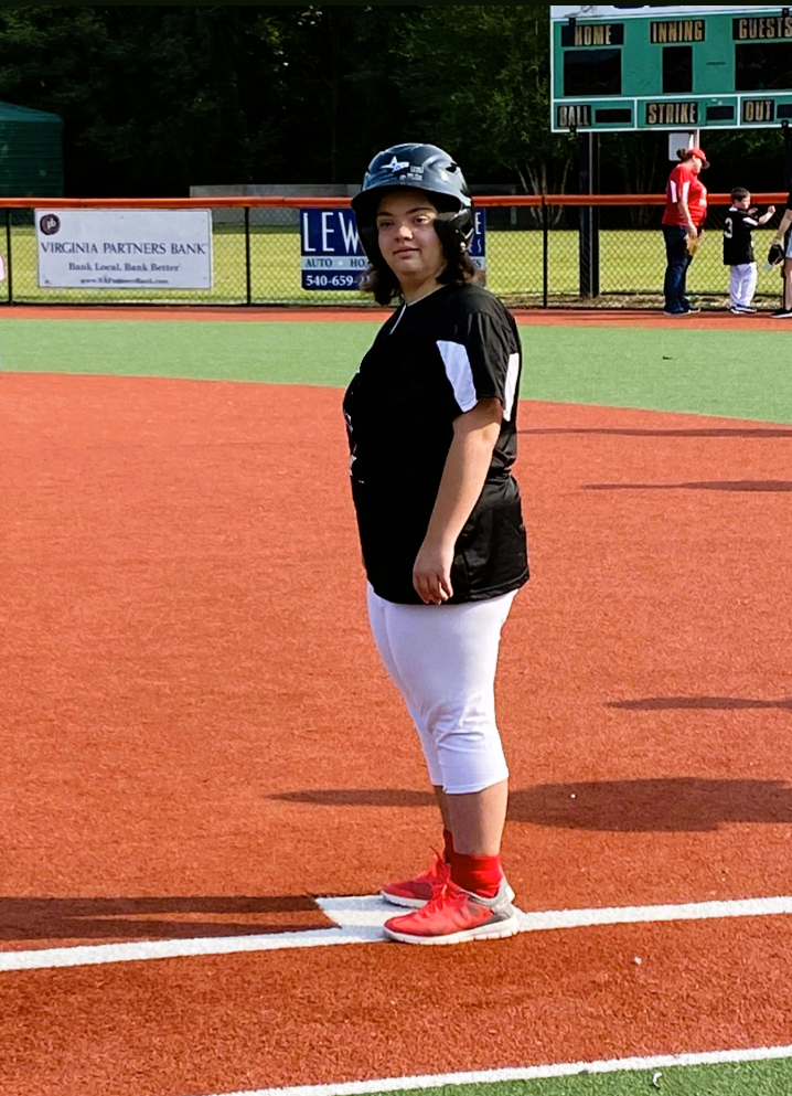 A young girl in softball uniform standing on a baseball field, wearing a helmet, black and white jersey, white pants, red socks, and red shoes.