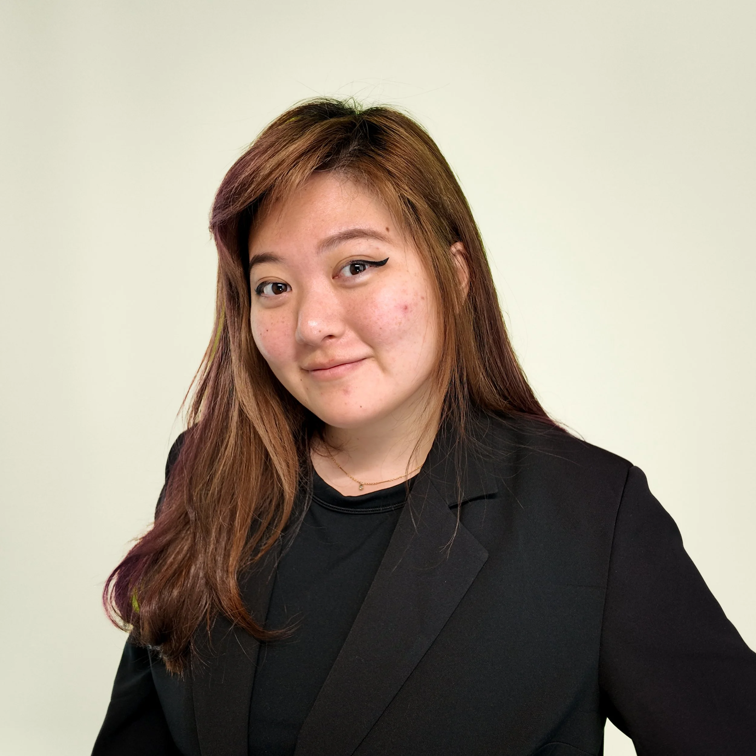 Partial view of a woman with long reddish-brown hair, black eyeliner, and freckles wearing a black top or blazer, standing against a light-colored background.