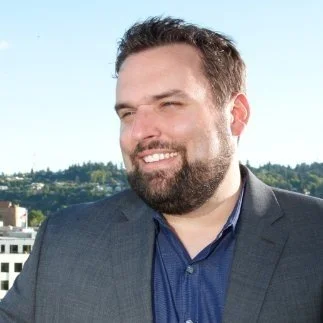 A man with a beard and short dark hair smiling outdoors, wearing a gray blazer and blue shirt, with a cityscape and trees in the background.