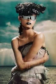 A woman with short, messy hair wearing a strapless top and skirt, standing against a cloudy sky background.