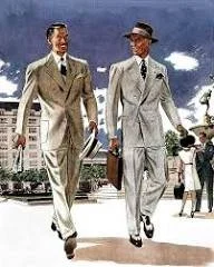Two men in suits walking outdoors on a city street with buildings and people in the background.