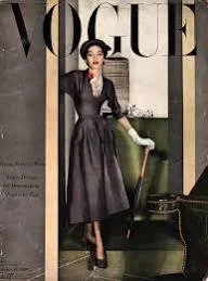 A woman in a black dress with a white collar stands in front of a Vogue magazine cover, holding a white clutch, with a green chair and patterned wall in the background.