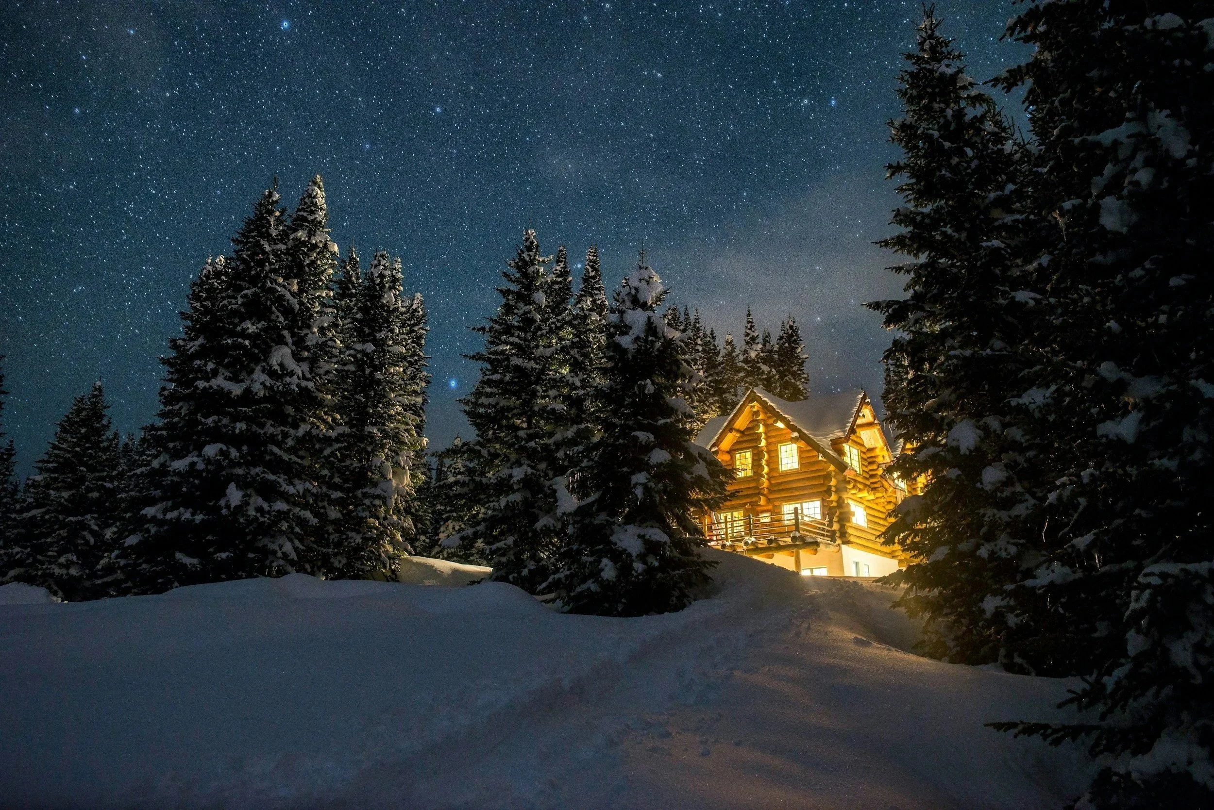 Snow-covered log cabin with lit windows surrounded by tall, snow-laden evergreen trees under a starry night sky.