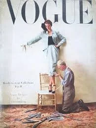 A woman standing on a chair in front of a Vogue magazine backdrop, being assisted by a man kneeling in front of her.