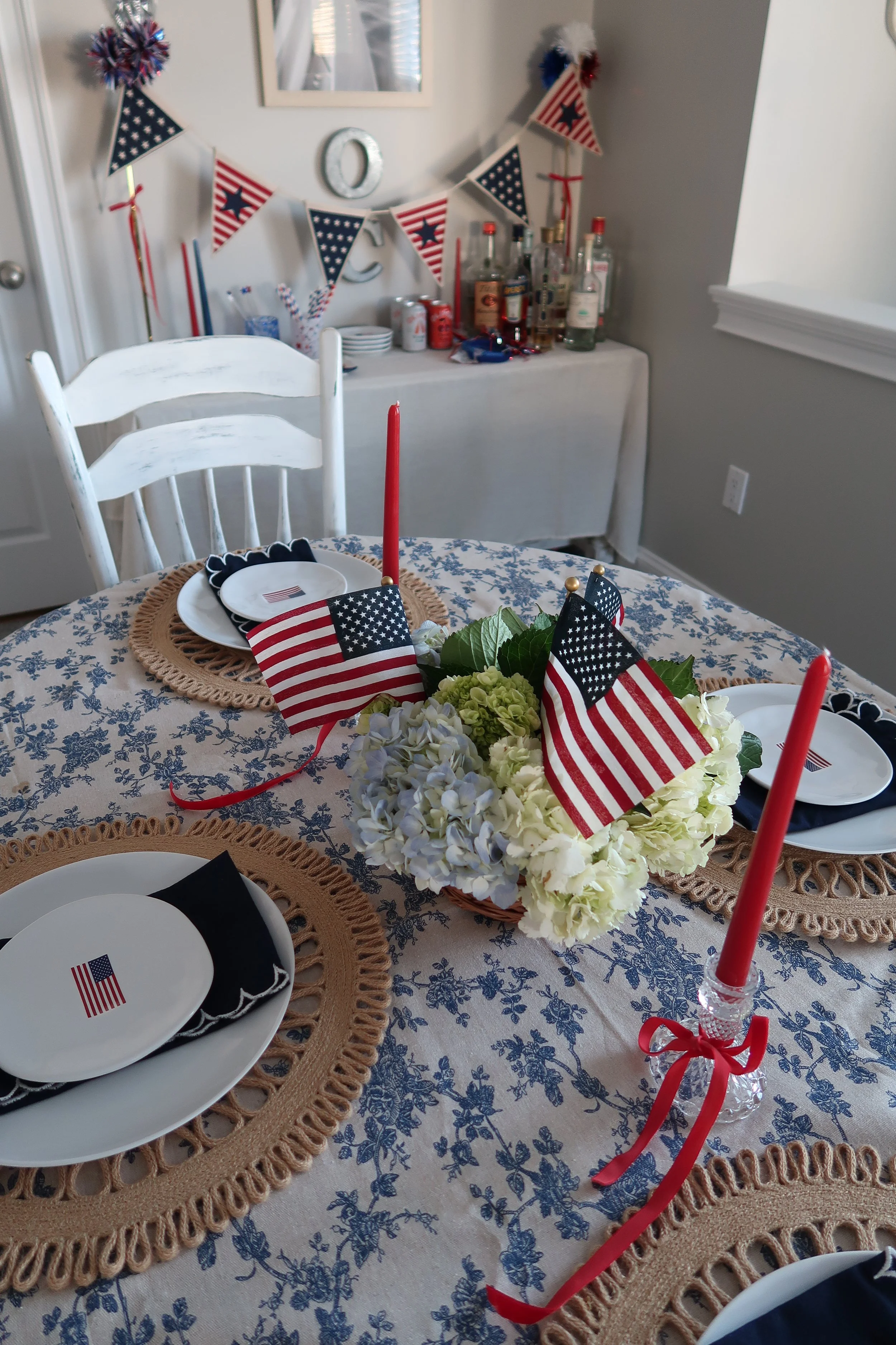 Table decorated for an American patriotic celebration with small American flags, red candles, and a floral centerpiece, with patriotic banners hanging on the wall in the background.