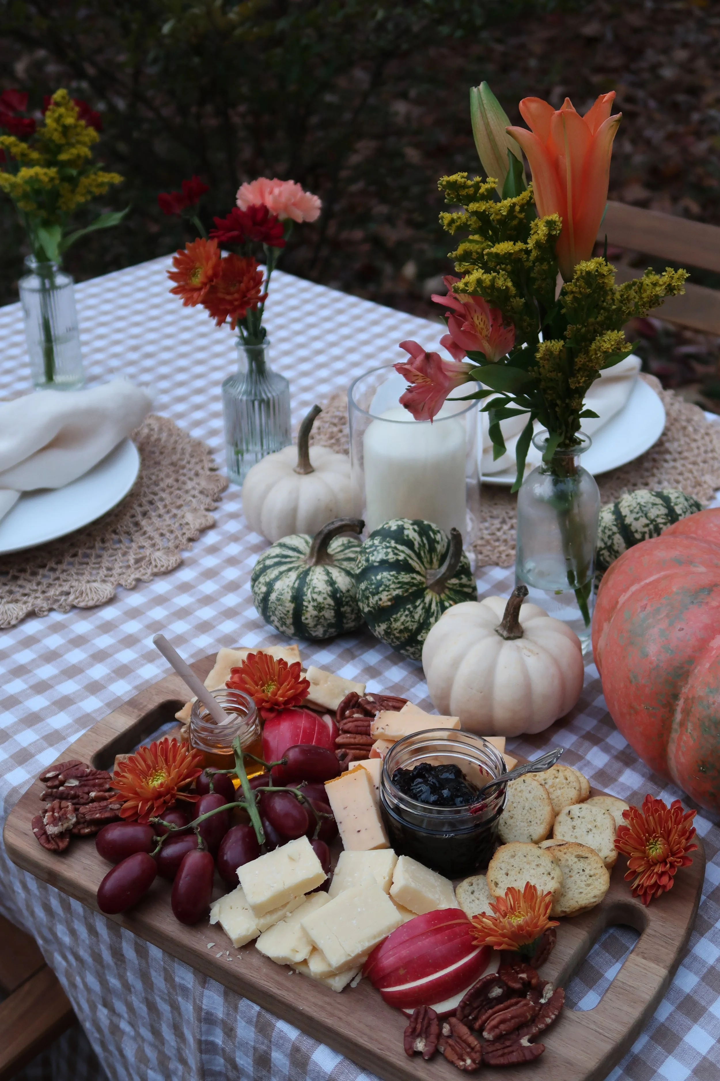 A fall-themed outdoor table setting with a cheese platter, grapes, and pumpkin decorations, topped with flower arrangements in vases.