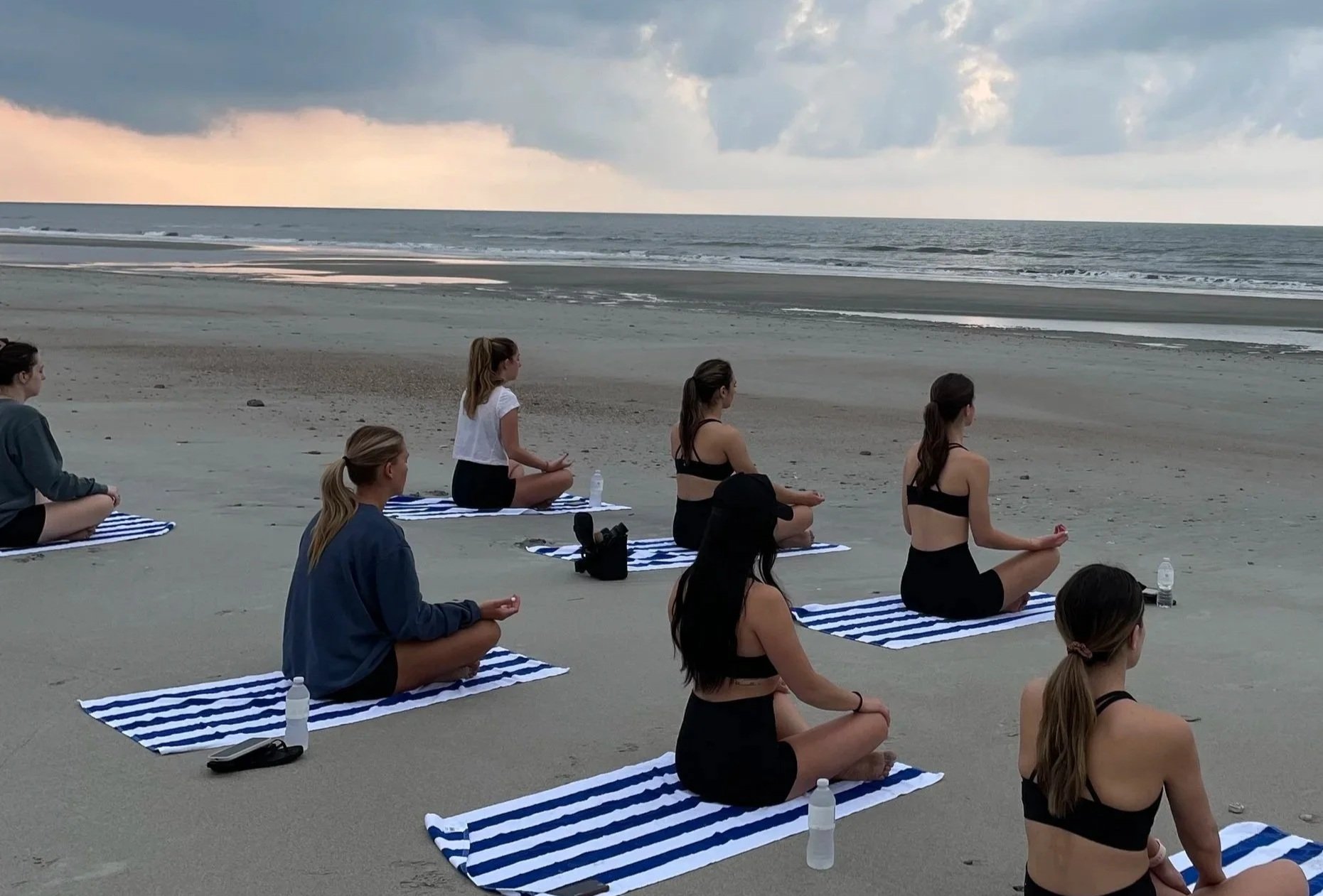 Group of women practicing yoga on the beach during sunset, sitting on blue and white striped mats facing the ocean.