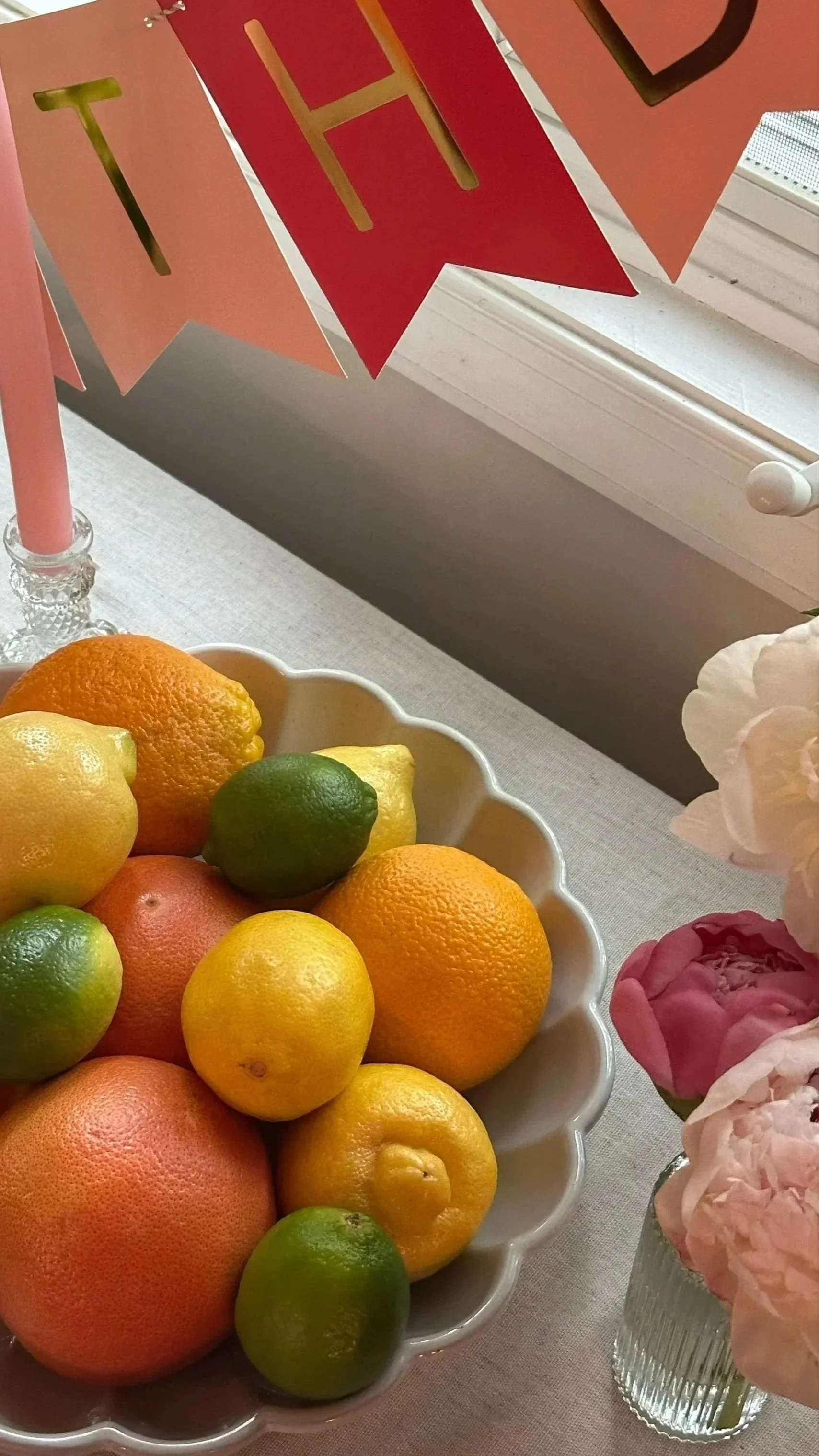 A bowl of assorted citrus fruits including lemons, oranges, and limes on a table, with pink flowers in a glass vase and a pink banner with gold lettering hanging above.