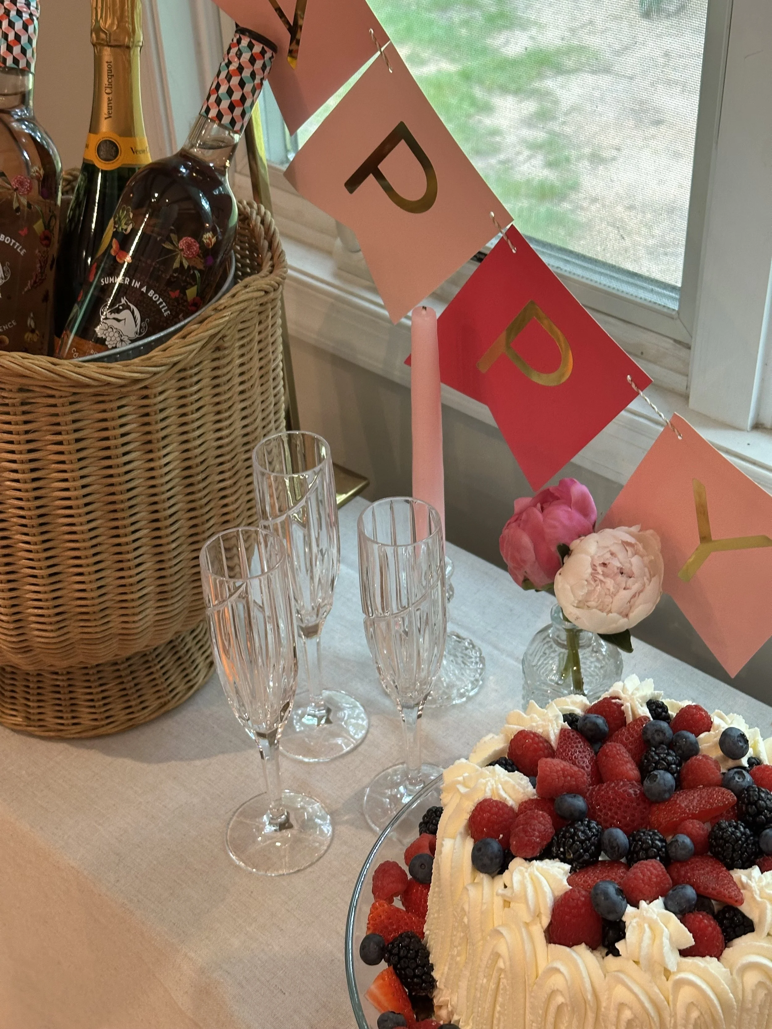 A celebration table with a fruit cake topped with berries, four empty champagne glasses, a pink candle, pink and white flowers, a pink "Happy Day" banner, a basket of bottles, and a window in the background.