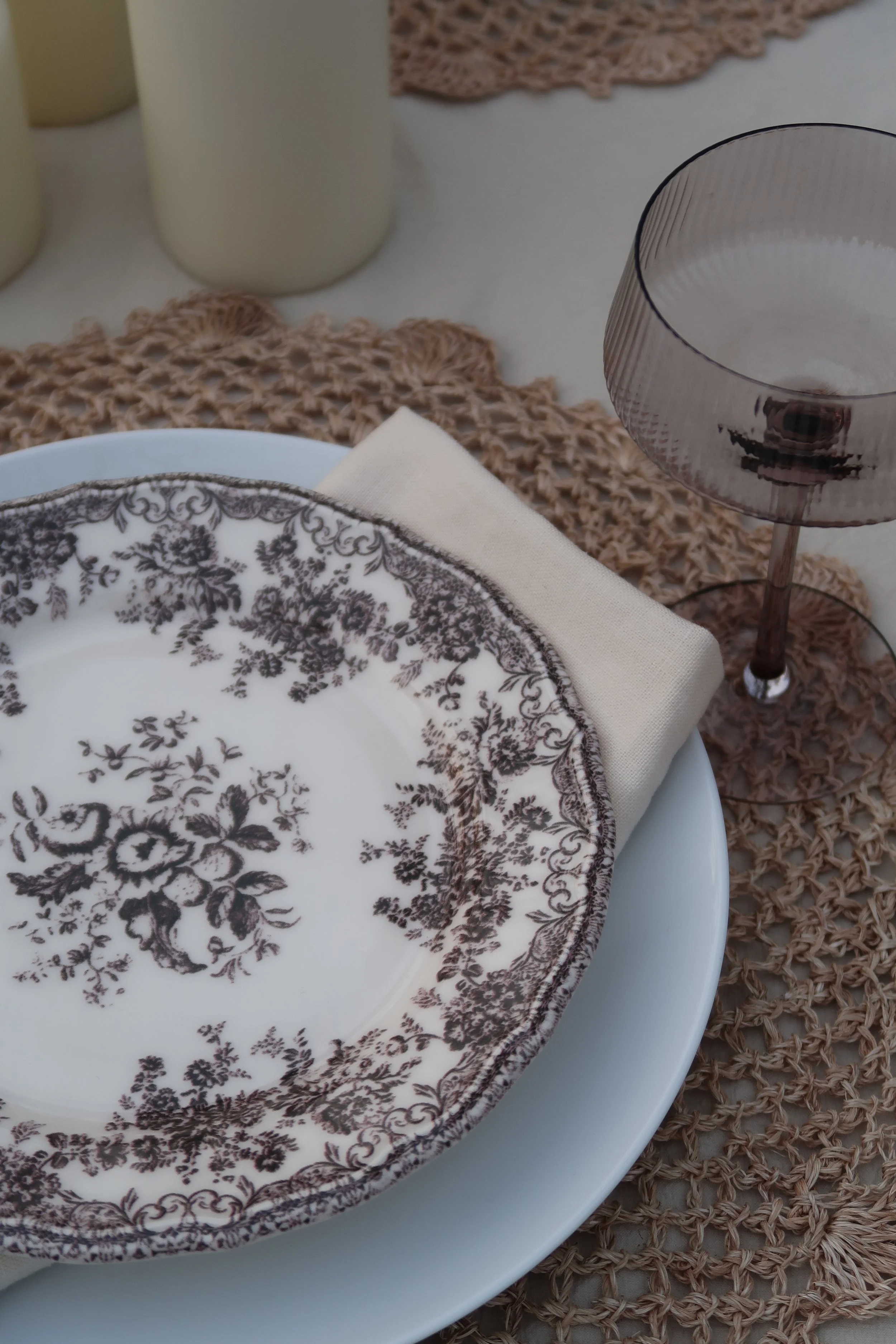 A table setting with a white plate featuring a dark floral pattern, a beige napkin, a clear purple-tinted wine glass, and multiple beige pillar candles on a woven placemat.