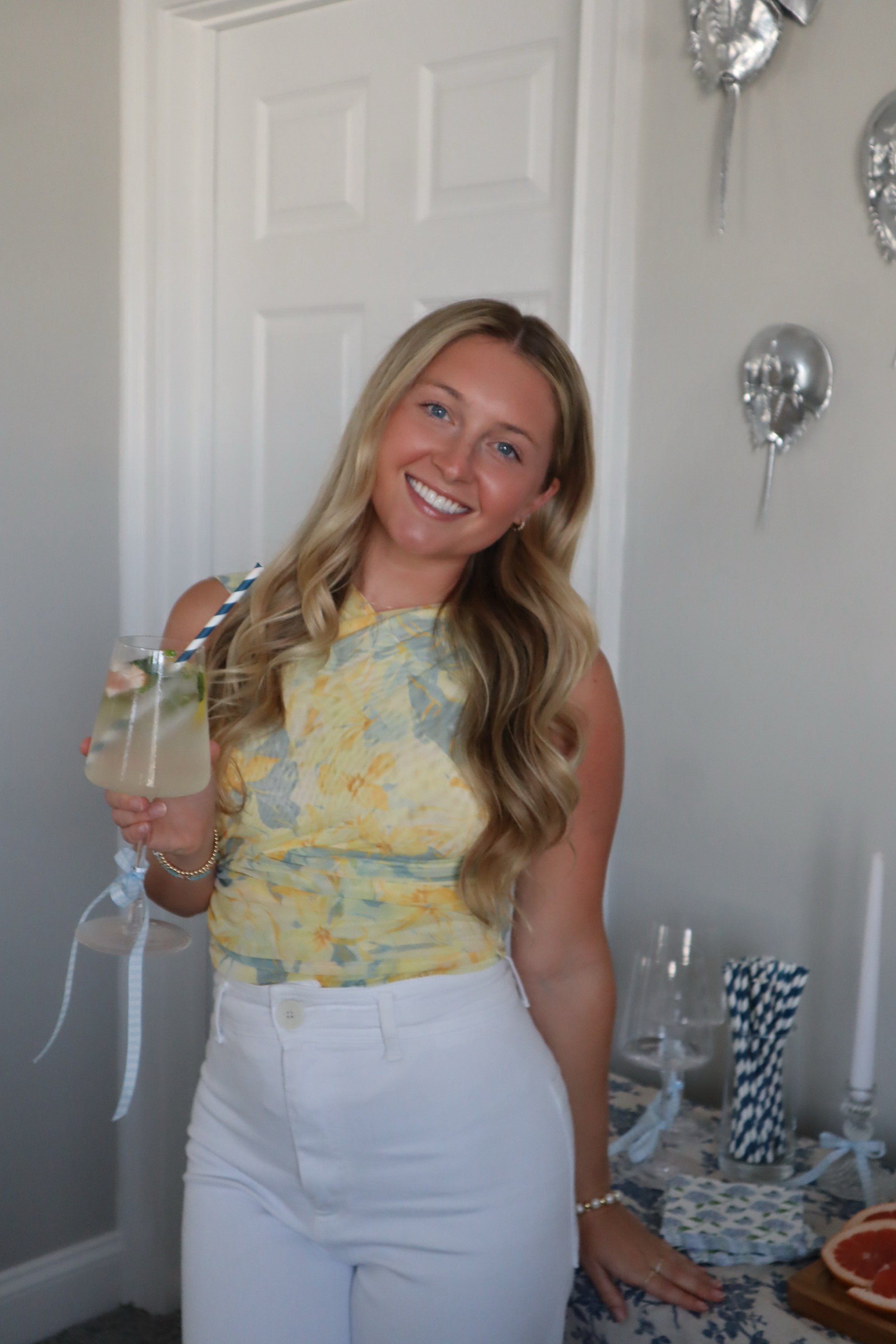 A woman with long blonde hair, wearing a yellow sleeveless top and white pants, smiling and holding a cocktail in her right hand. She is standing near a decorated table with glasses, napkins, and a grapefruit slice in the background.