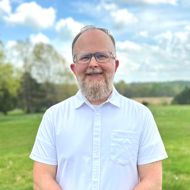 A smiling man with glasses and a beard in a white short-sleeve button-up shirt standing outdoors in a grassy field with trees and a cloudy sky in the background.