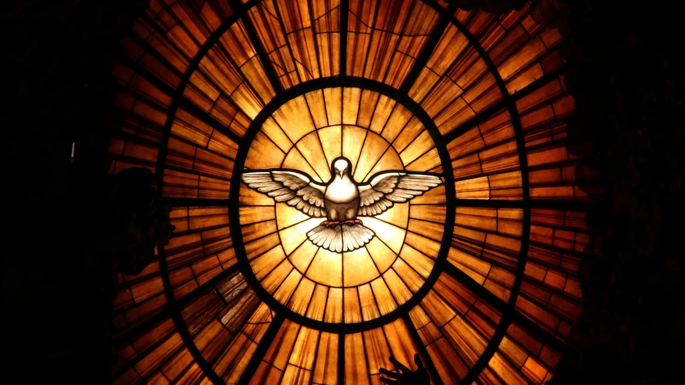 A stained glass window depicting a dove with outstretched wings, surrounded by orange and brown geometric patterns.