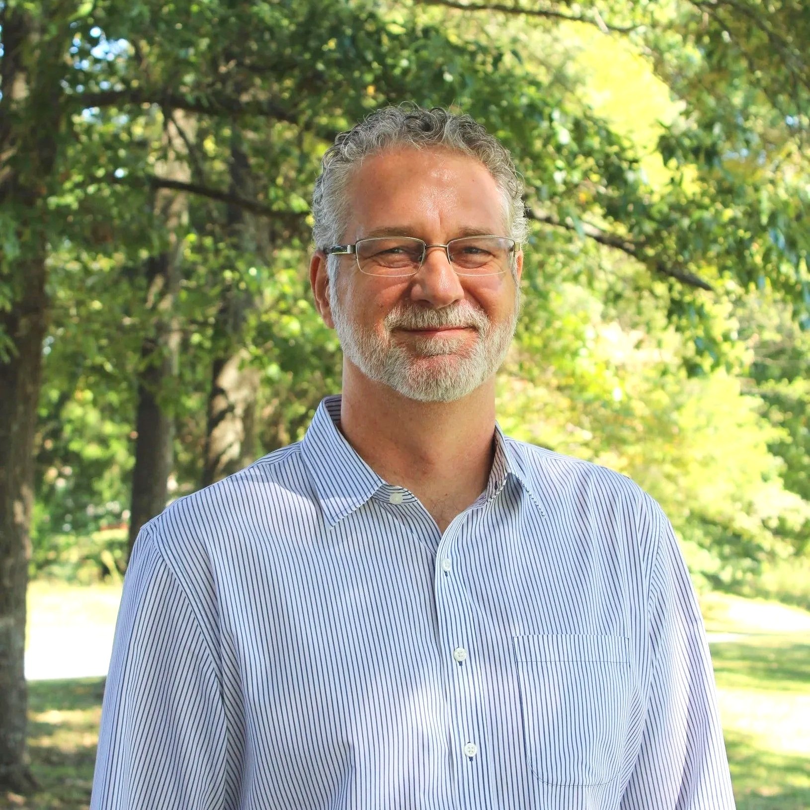 A smiling middle-aged man with gray hair, beard, and glasses standing outdoors with lush green trees and sunlight in the background.