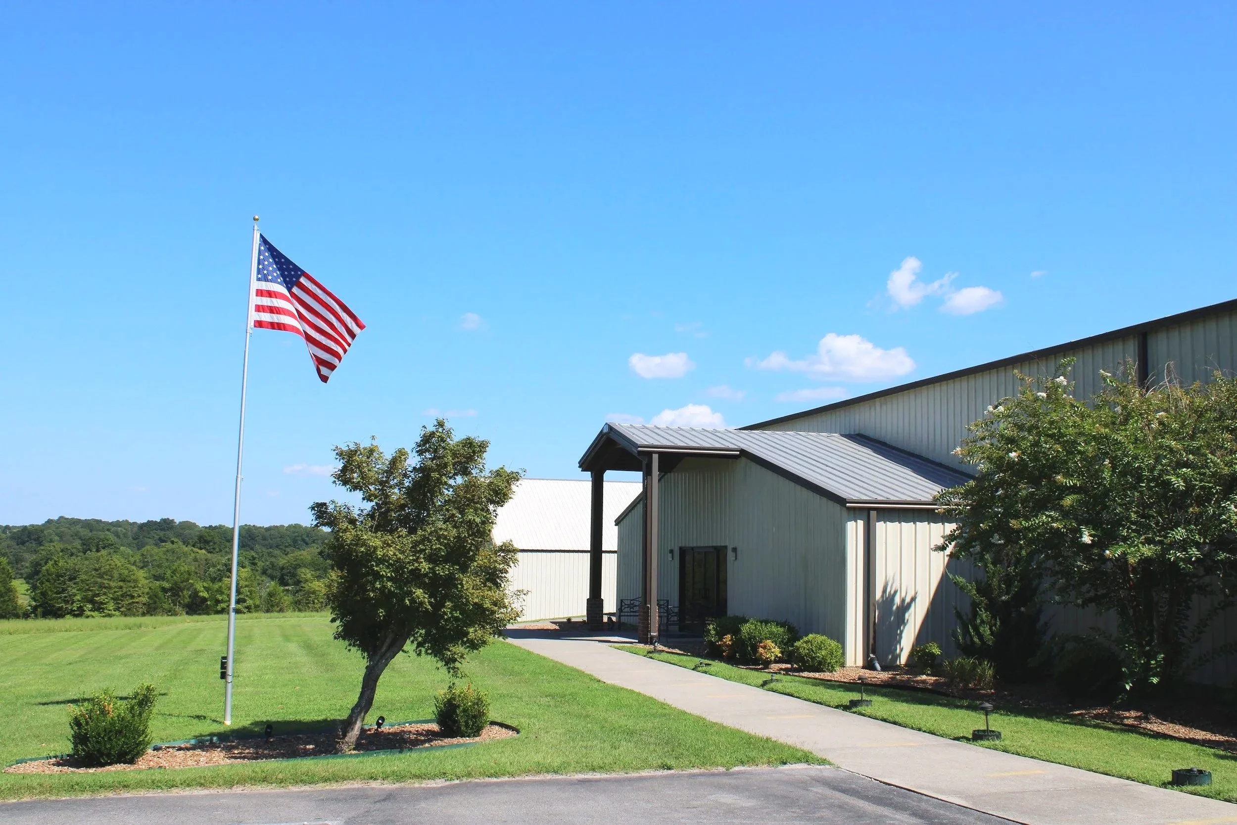 A blue sky with a few white clouds, an American flag on a flagpole, a green lawn, a tree, and a beige building with a metal roof and a sidewalk in front.
