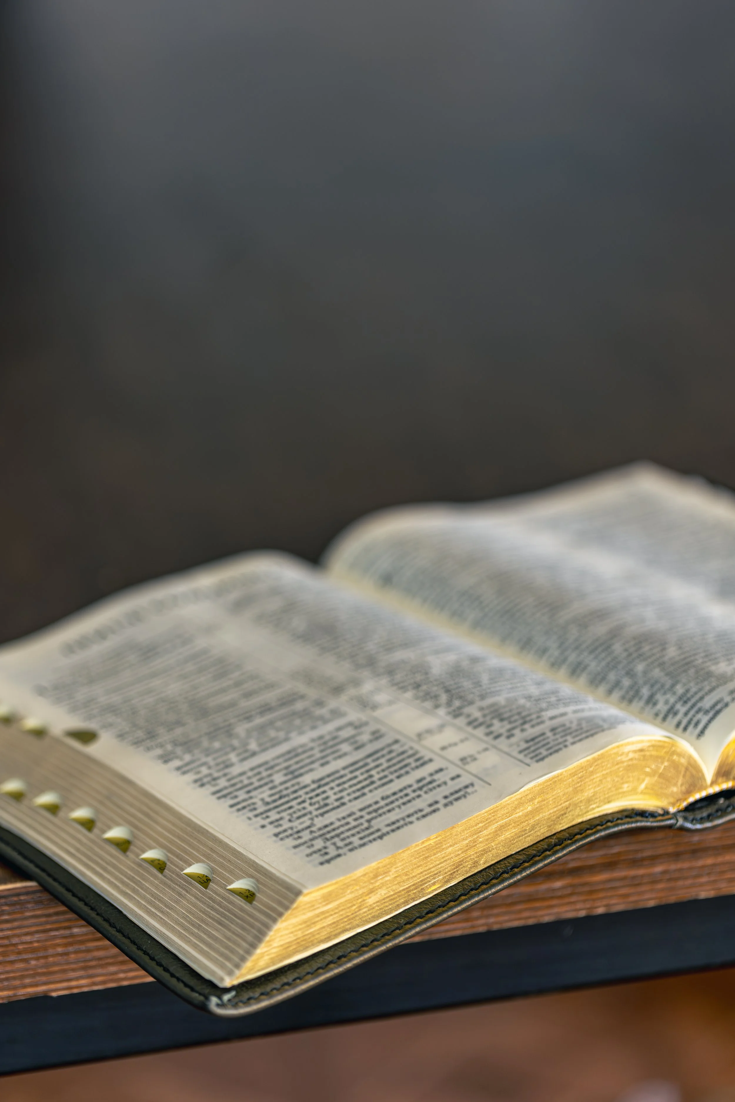 An open Bible with yellowed pages resting on a wooden surface, with a dark, blurred background.