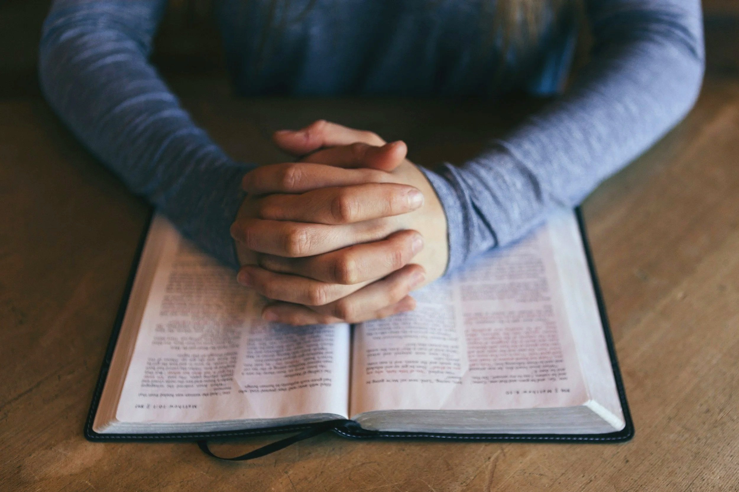 Person sitting at a table with hands clasped over an open Bible.