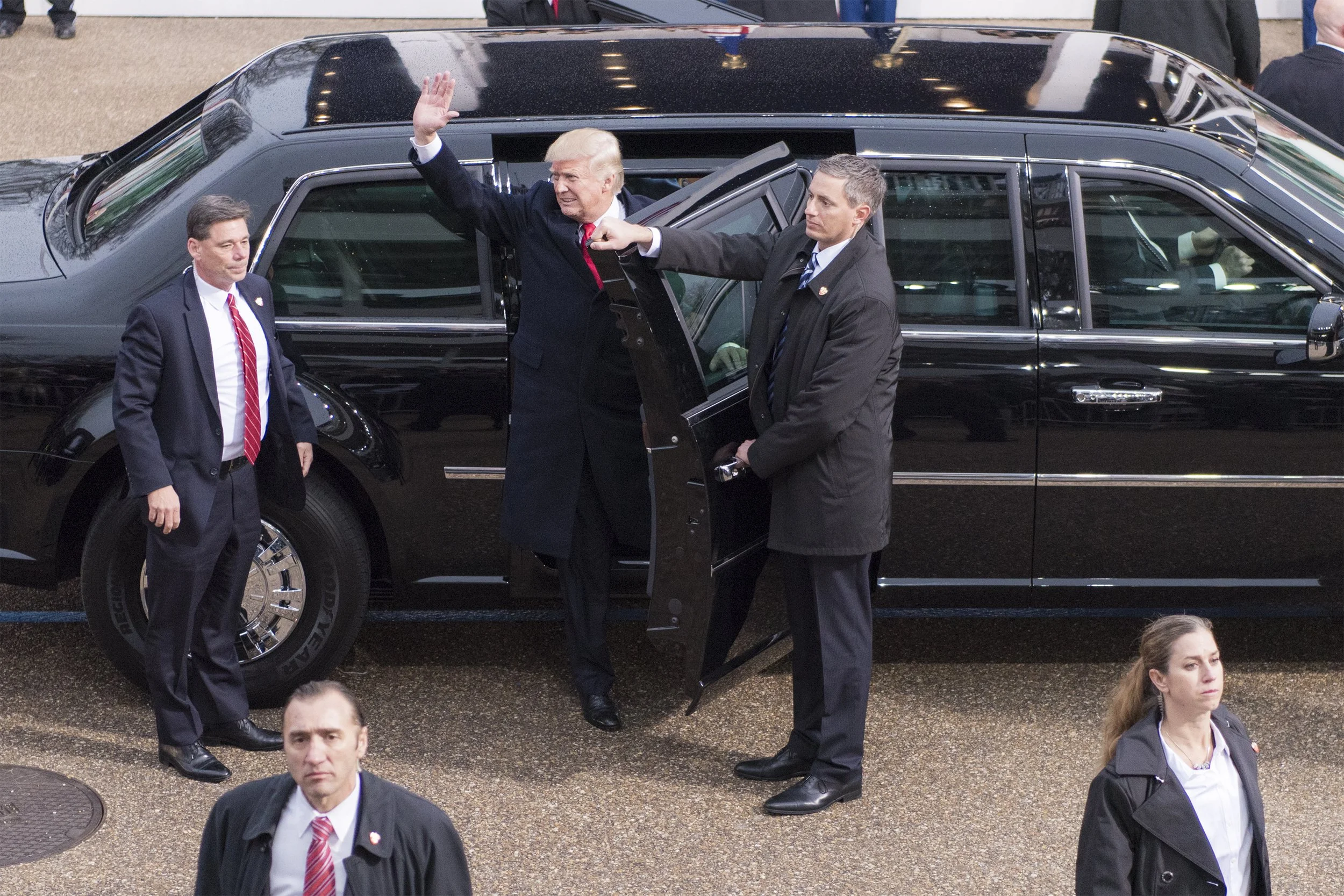 President Donald Trump waves while standing in a black limousine, accompanied by security personnel.