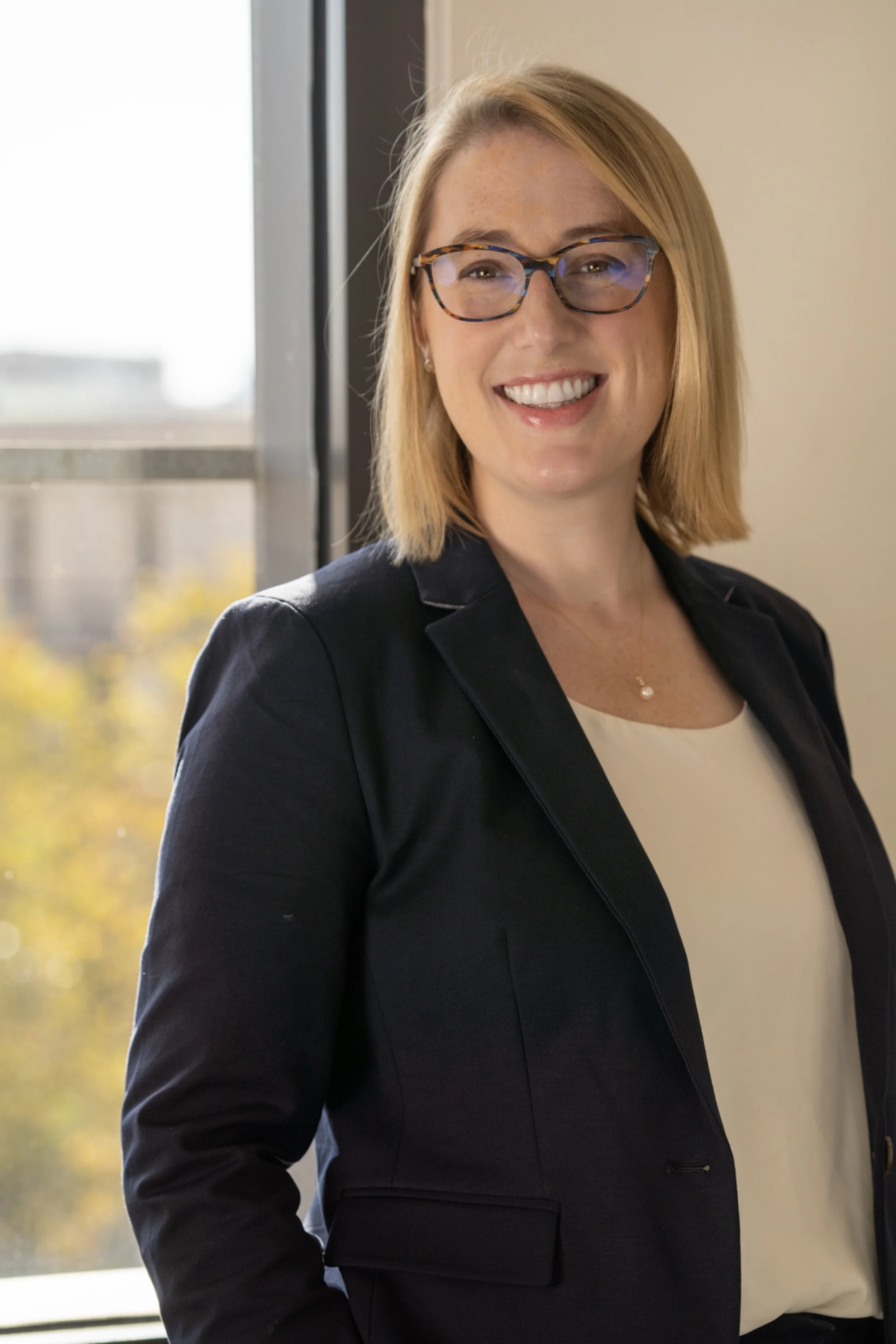 A woman with blonde hair, wearing glasses, a black blazer, and a white top, smiling while standing indoors near a window with cityscape and trees in the background.