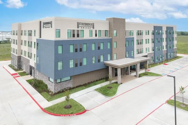 Modern Staybridge Suites by IHG with a covered entrance, situated in a suburban area with green fields in the background.