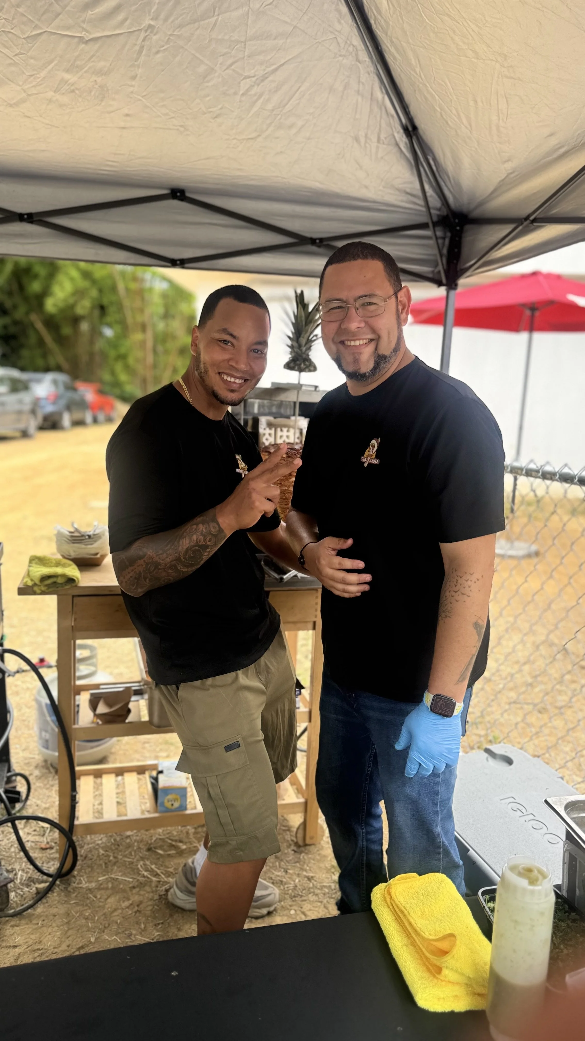 Dos hombres sonrientes en un puesto de comida al aire libre, uno con guantes azules y ambos con camisetas negras.