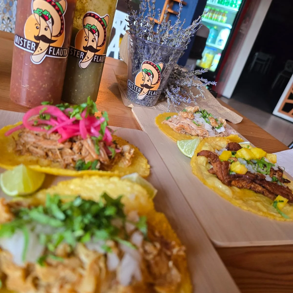 Tacos con diferentes ingredientes como carne, pollo, y vegetales en una mesa de madera, con bebidas en botellas cómico-recortadas y una maceta con plantas decorativas.