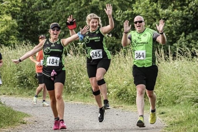 Three runners in green and black athletic gear waving and smiling while running outdoors on a grassy trail.