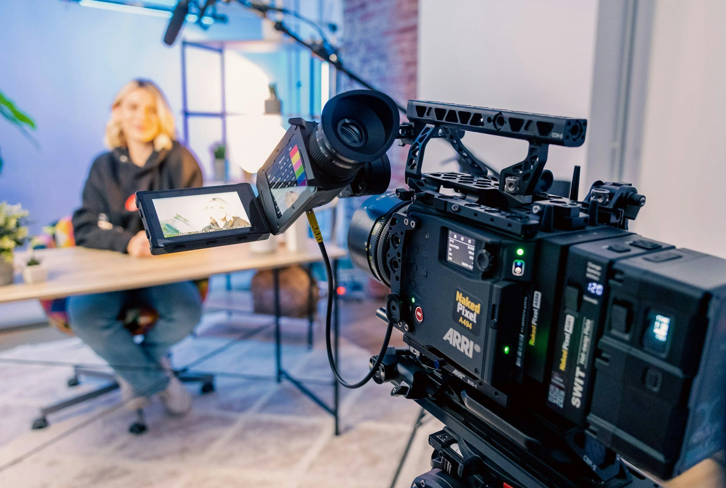Filming equipment, including a professional camera and monitor, set up in a room where a woman in casual attire is sitting at a table with plants and decor, appearing in focus on the camera.