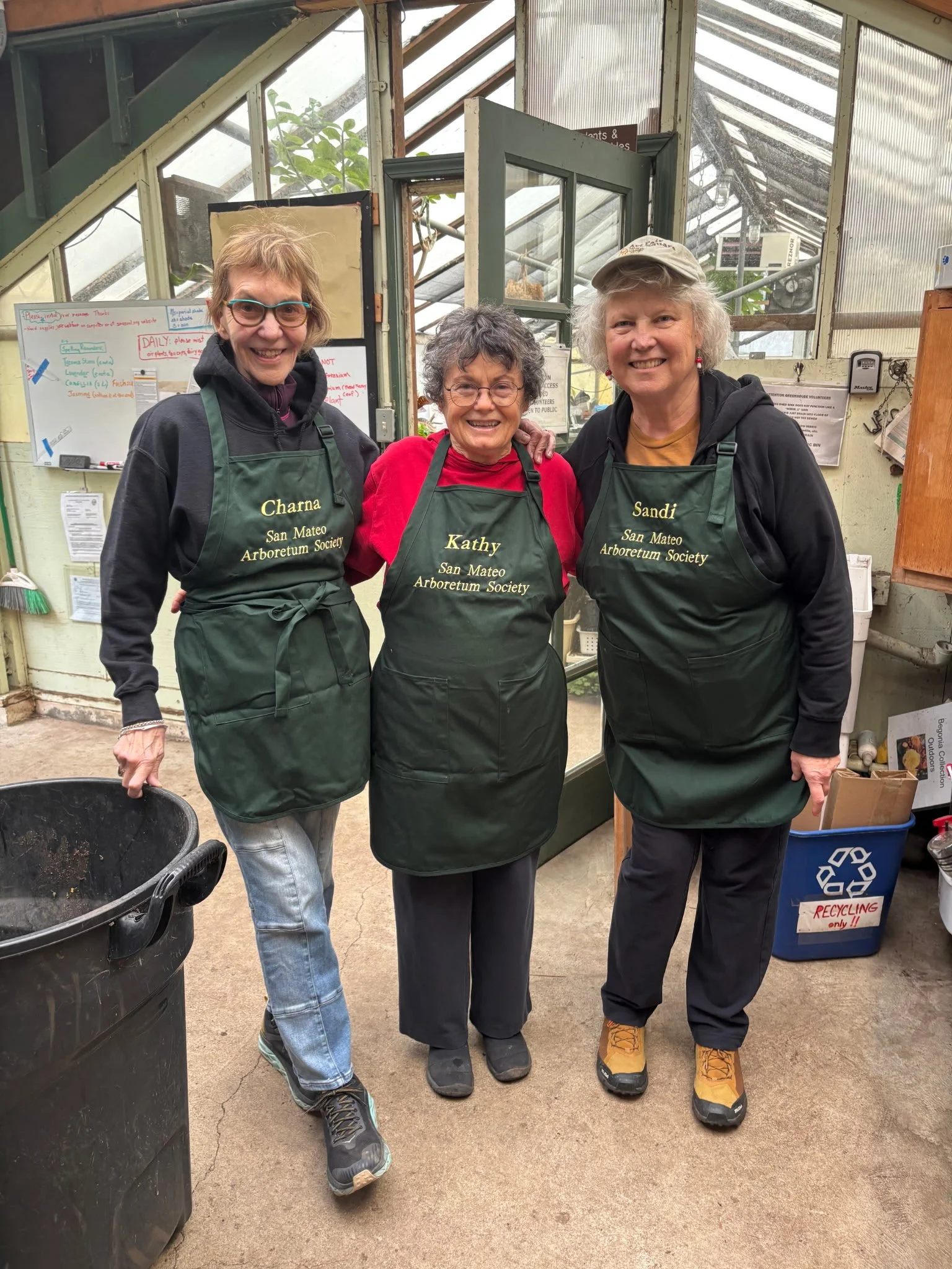 Three women wearing green aprons with names and 'San Mateo Arboretum Society' standing inside a greenhouse, smiling.