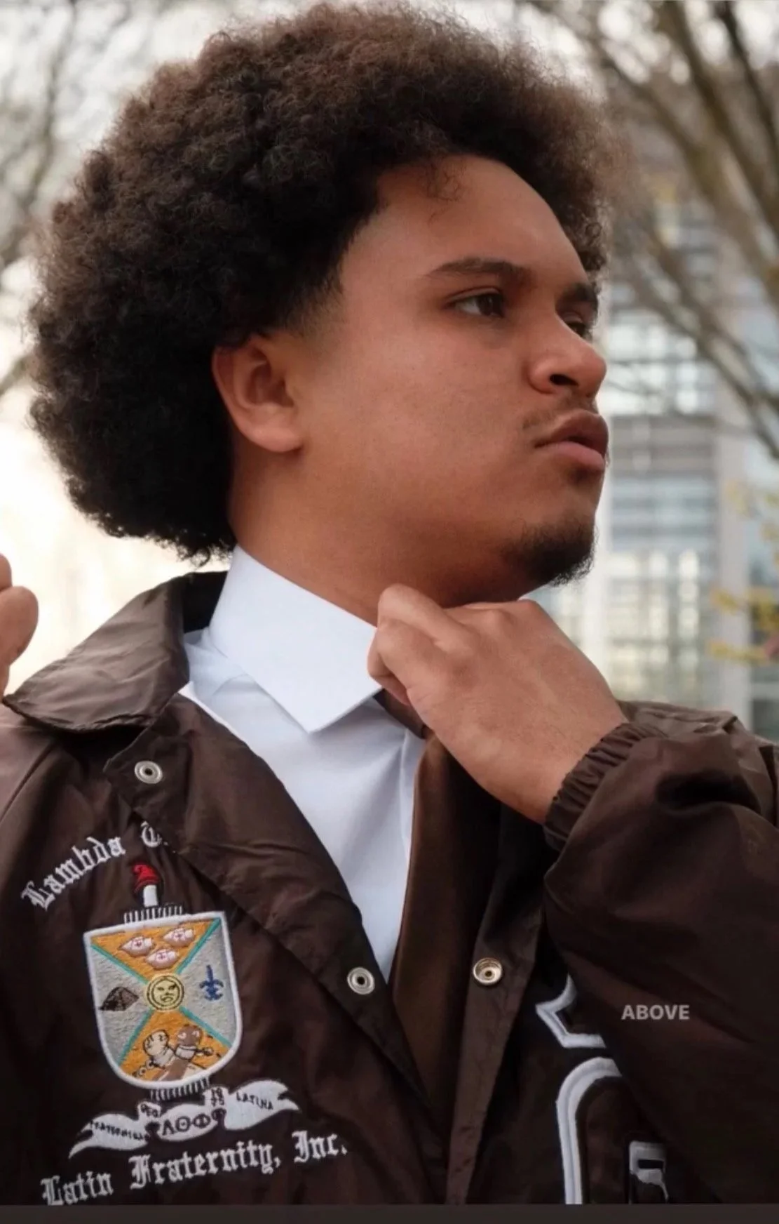A young man with curly hair adjusting his collar outdoors, wearing a brown jacket with patches and text.