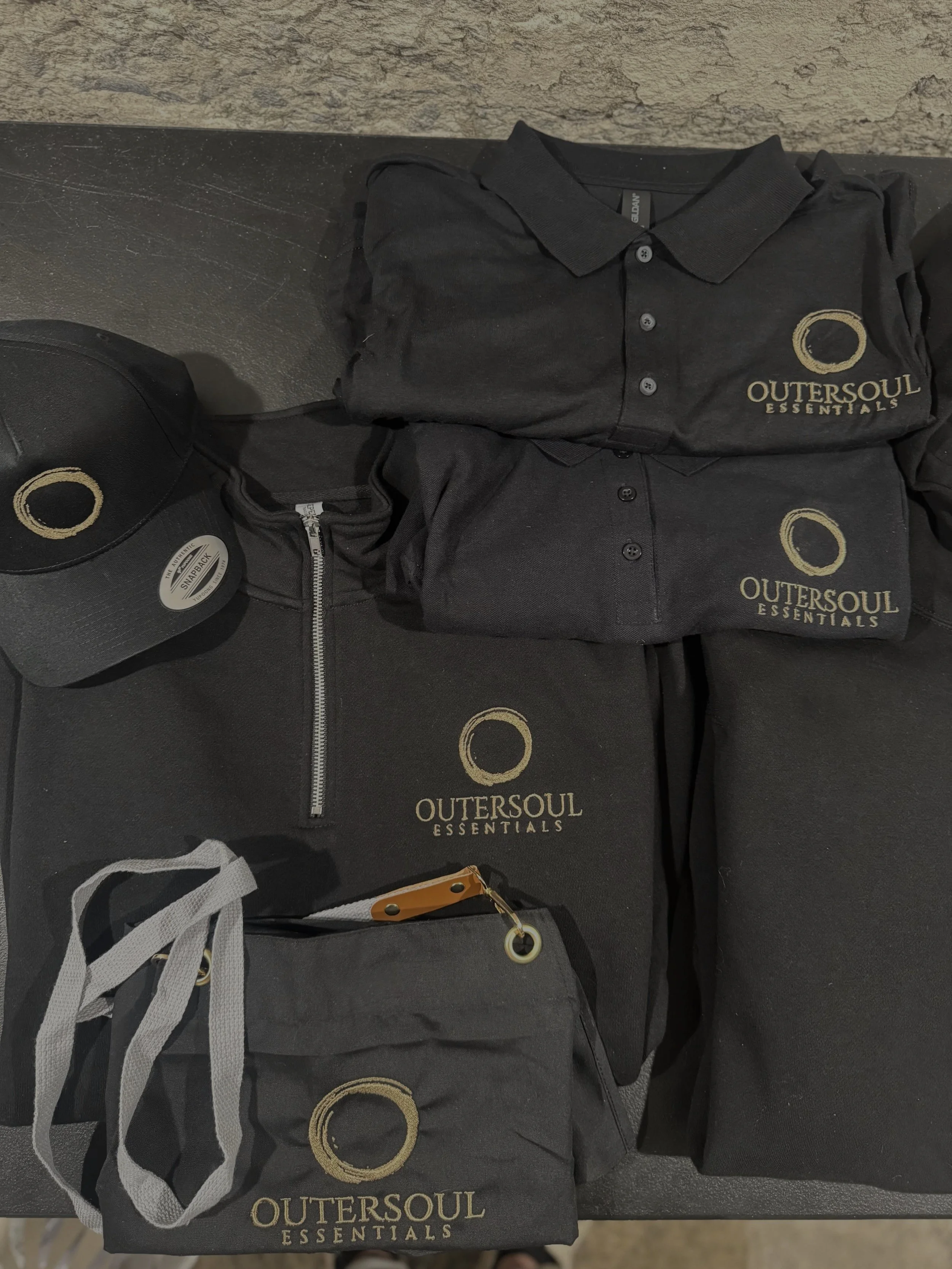 Various black clothing and accessories with Outersoul Essentials logo, including a hoodie with a zipper, a polo shirt, a cap, a small drawstring bag, and a larger bag, all placed on a black surface.